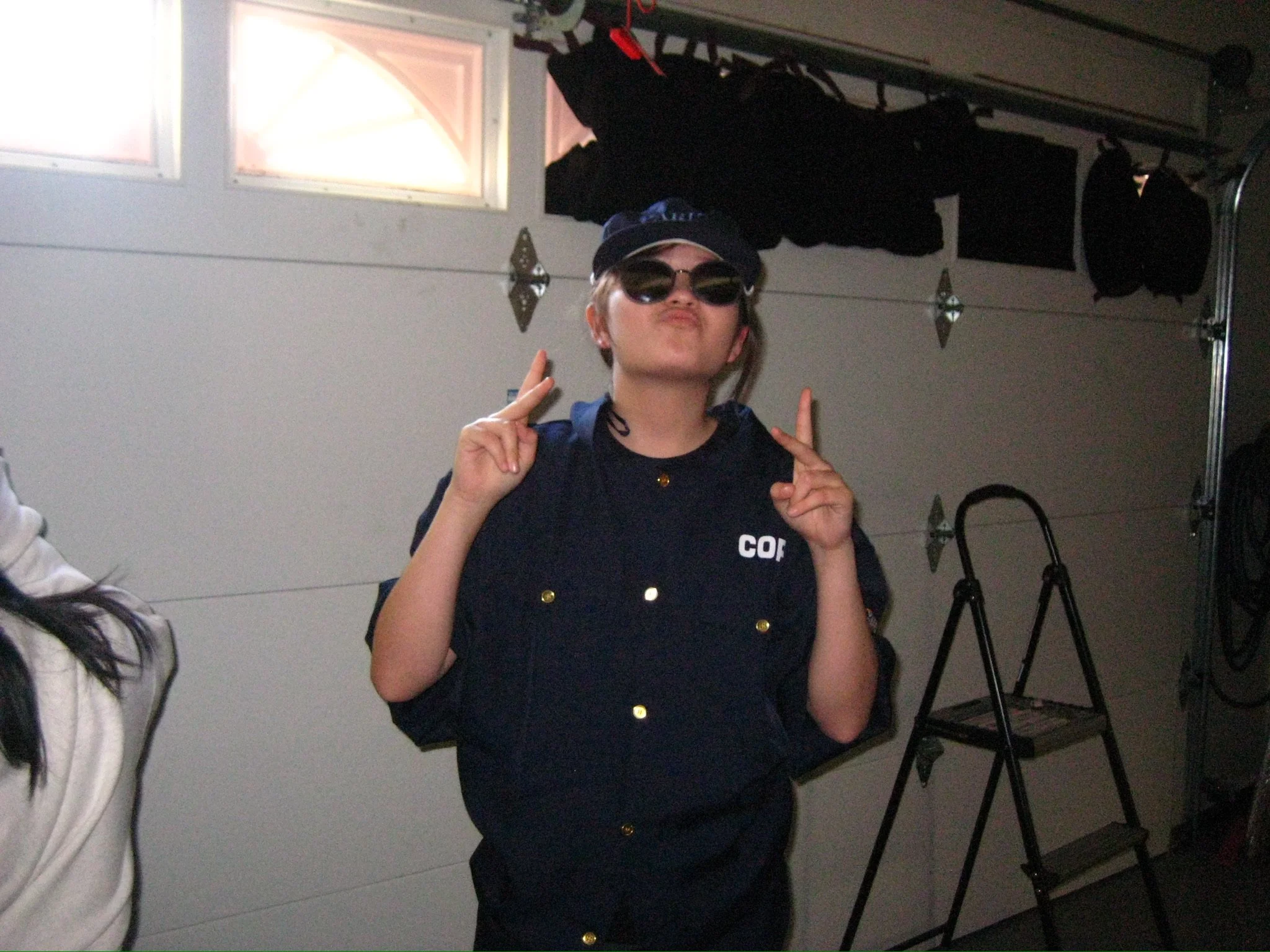 Person wearing sunglasses and a navy costume poses with both hands raised inside a garage.