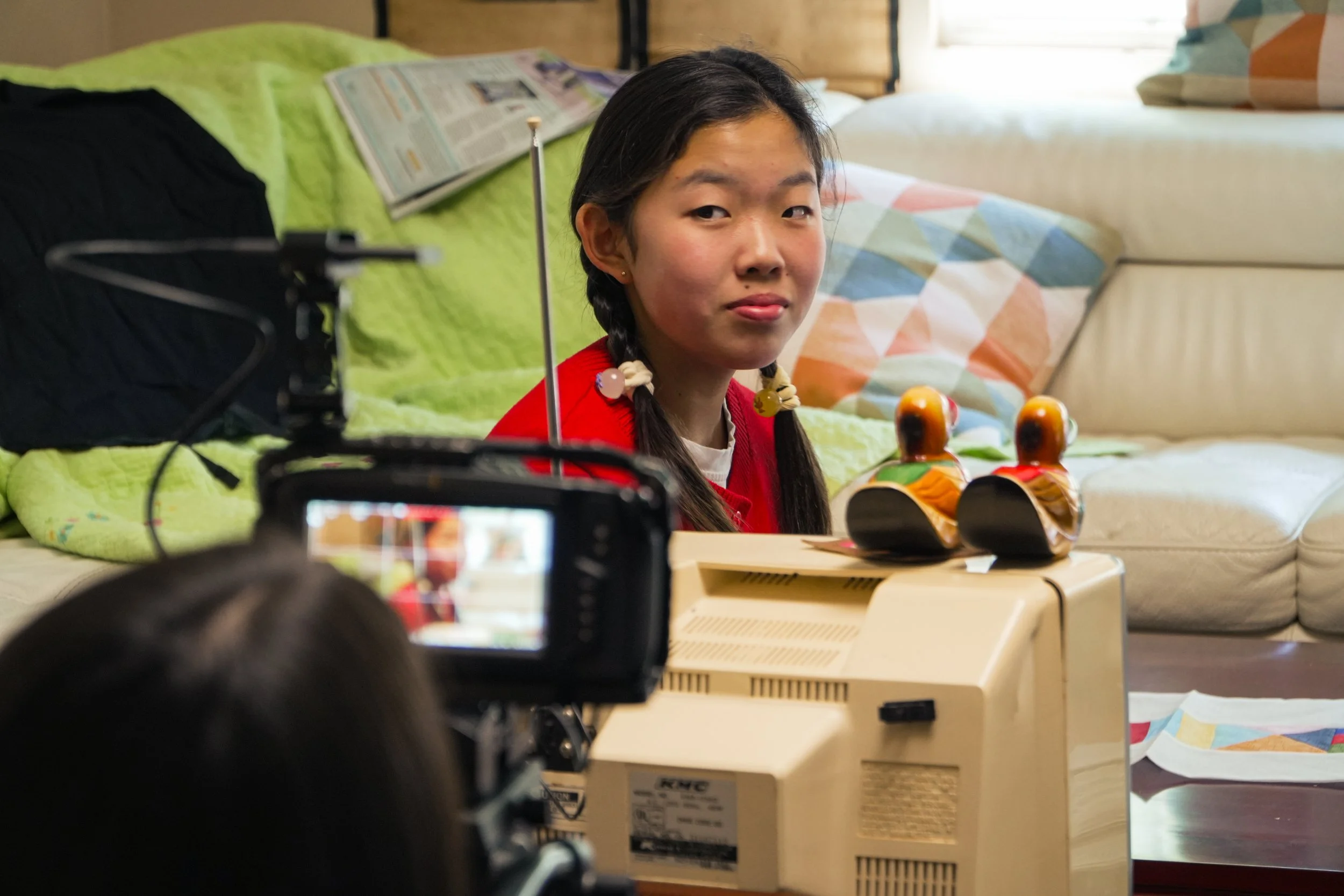 Young girl in a red outfit sits behind a vintage computer while being filmed in a bedroom.