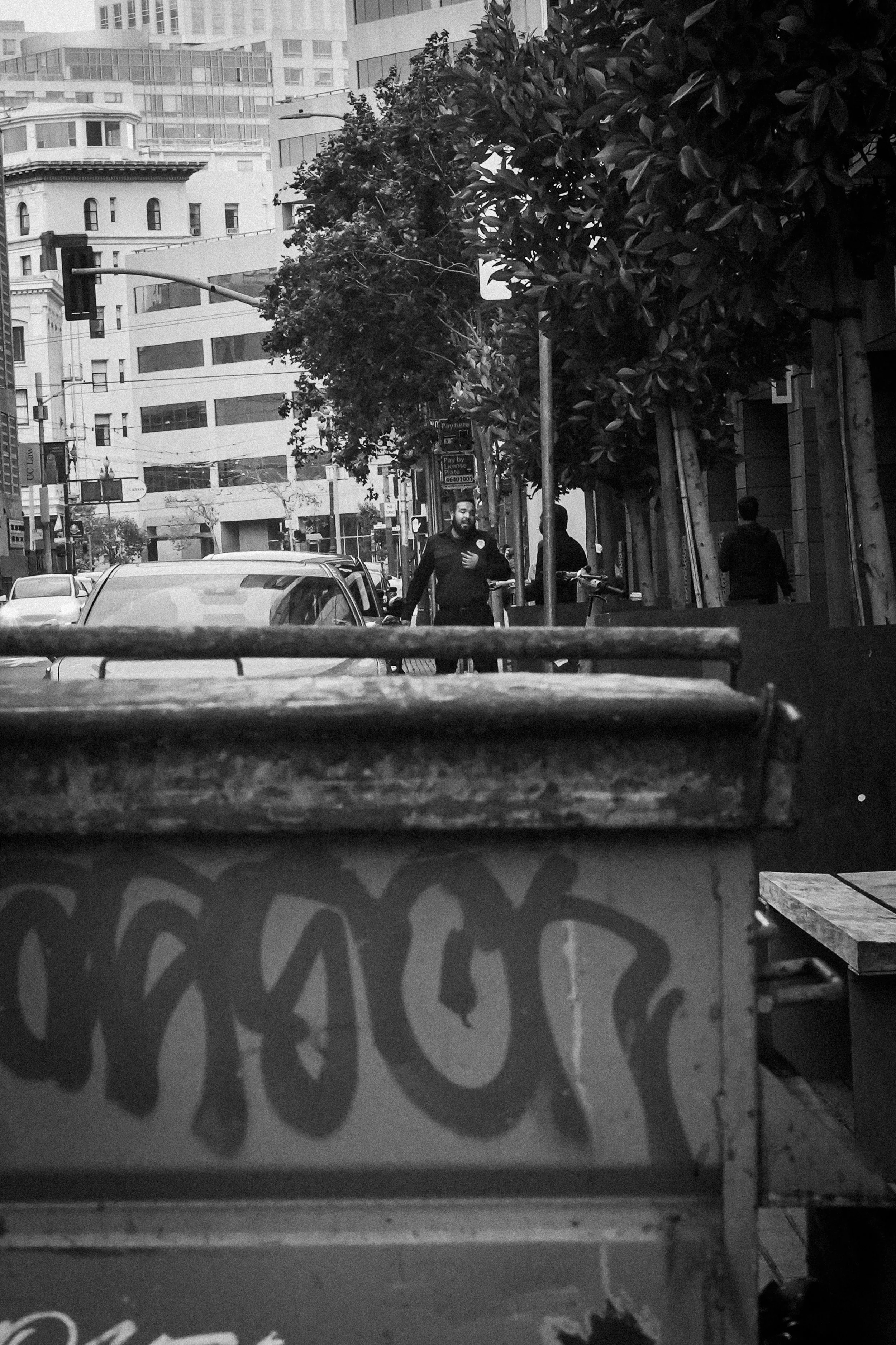 Black-and-white street scene with pedestrians, parked cars, traffic lights, leafy trees, and urban buildings viewed through a concrete barrier.