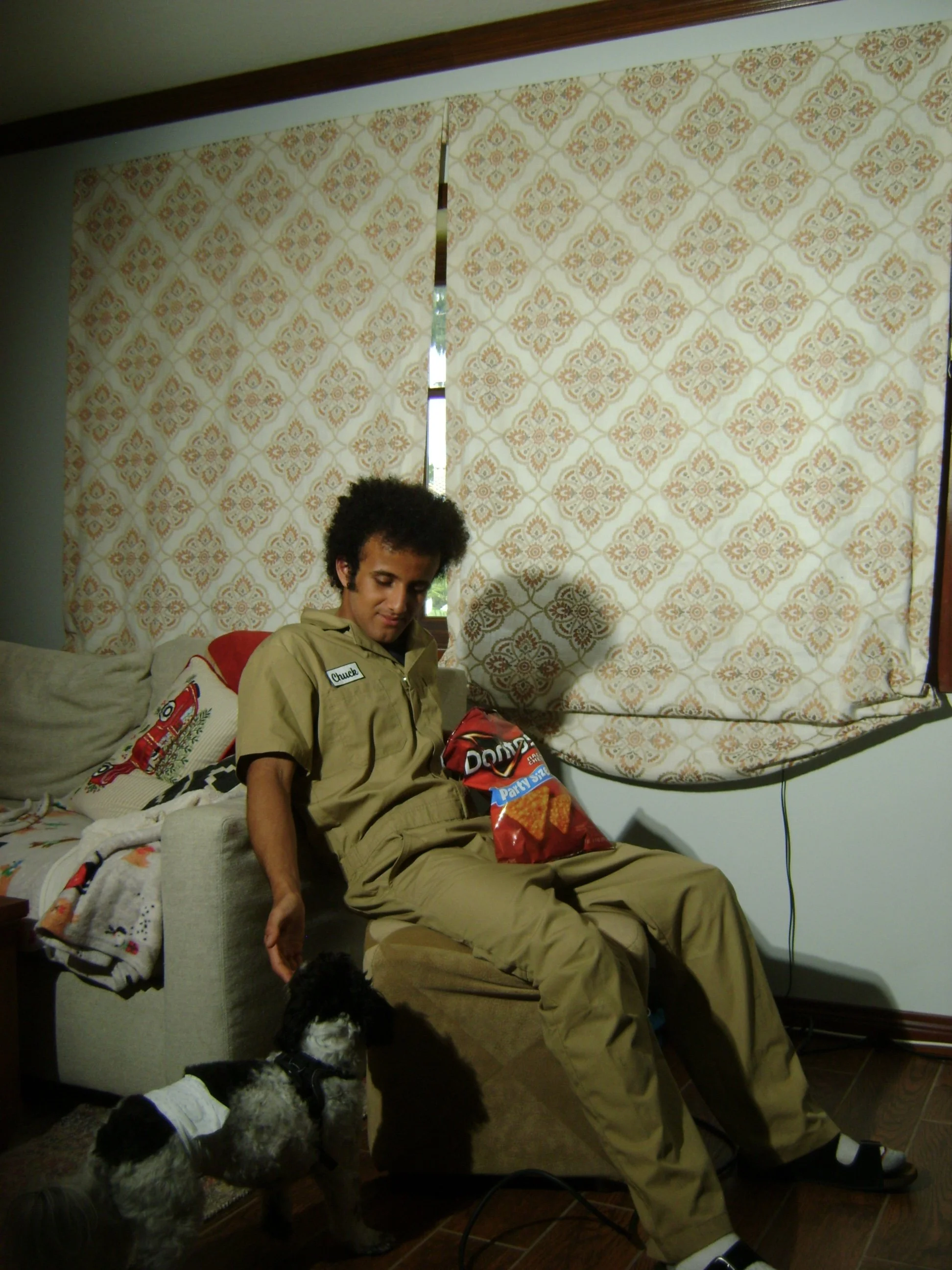 Young man sitting in a recliner with a bag of Doritos, while a small black-and-white dog rests on the floor nearby.