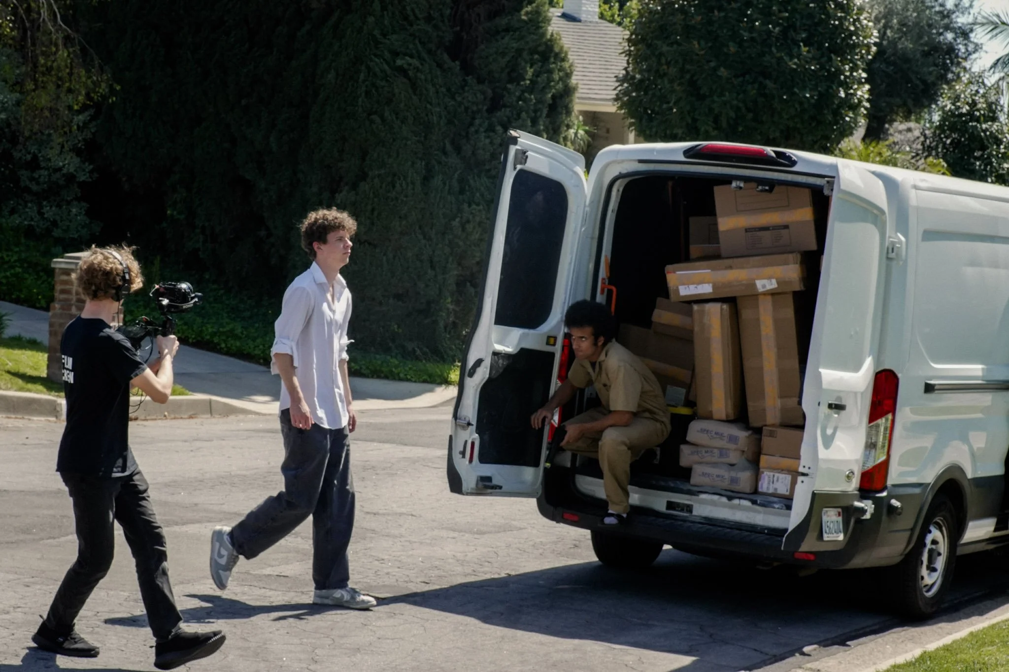 Film crew records a man loading cardboard boxes into a white delivery van on a suburban street