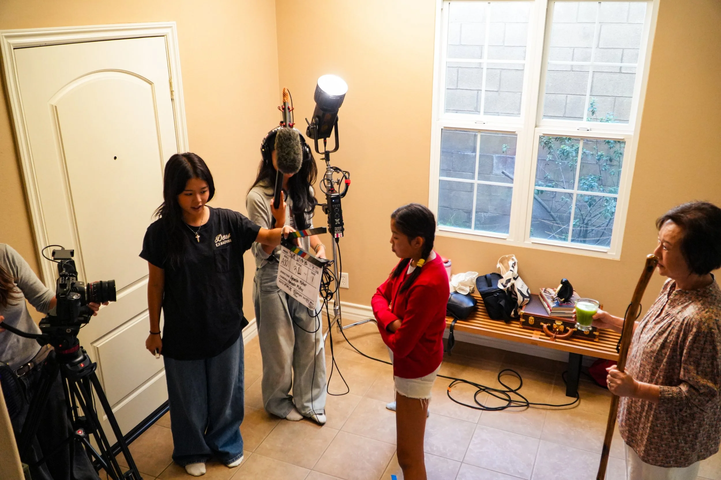 Film crew recording a scene indoors with cameras, boom microphone, and clapperboard while actors and crew members gather nearby