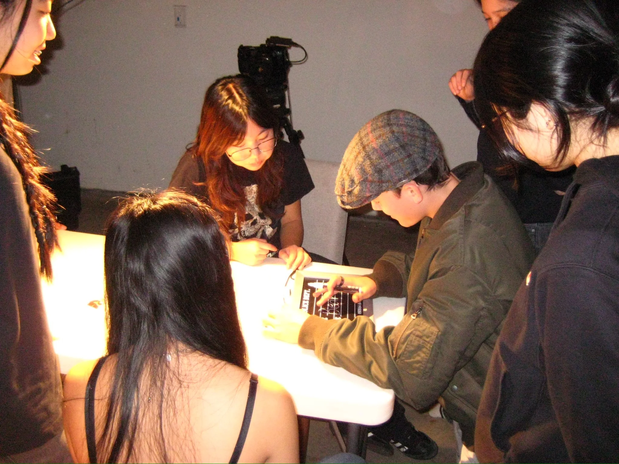Group of people gathered around a table, with one person writing in a notebook and another recording with a video camera.