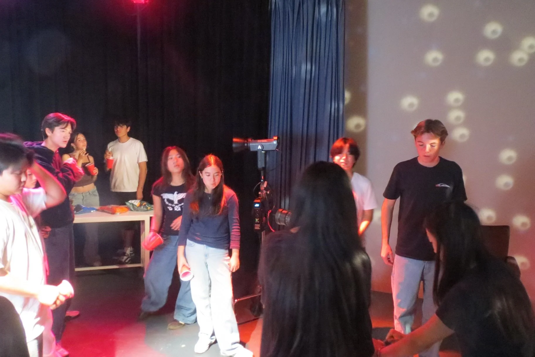 Group of young people gathered on a stage or event set under colorful lighting, with a camera filming in the foreground.