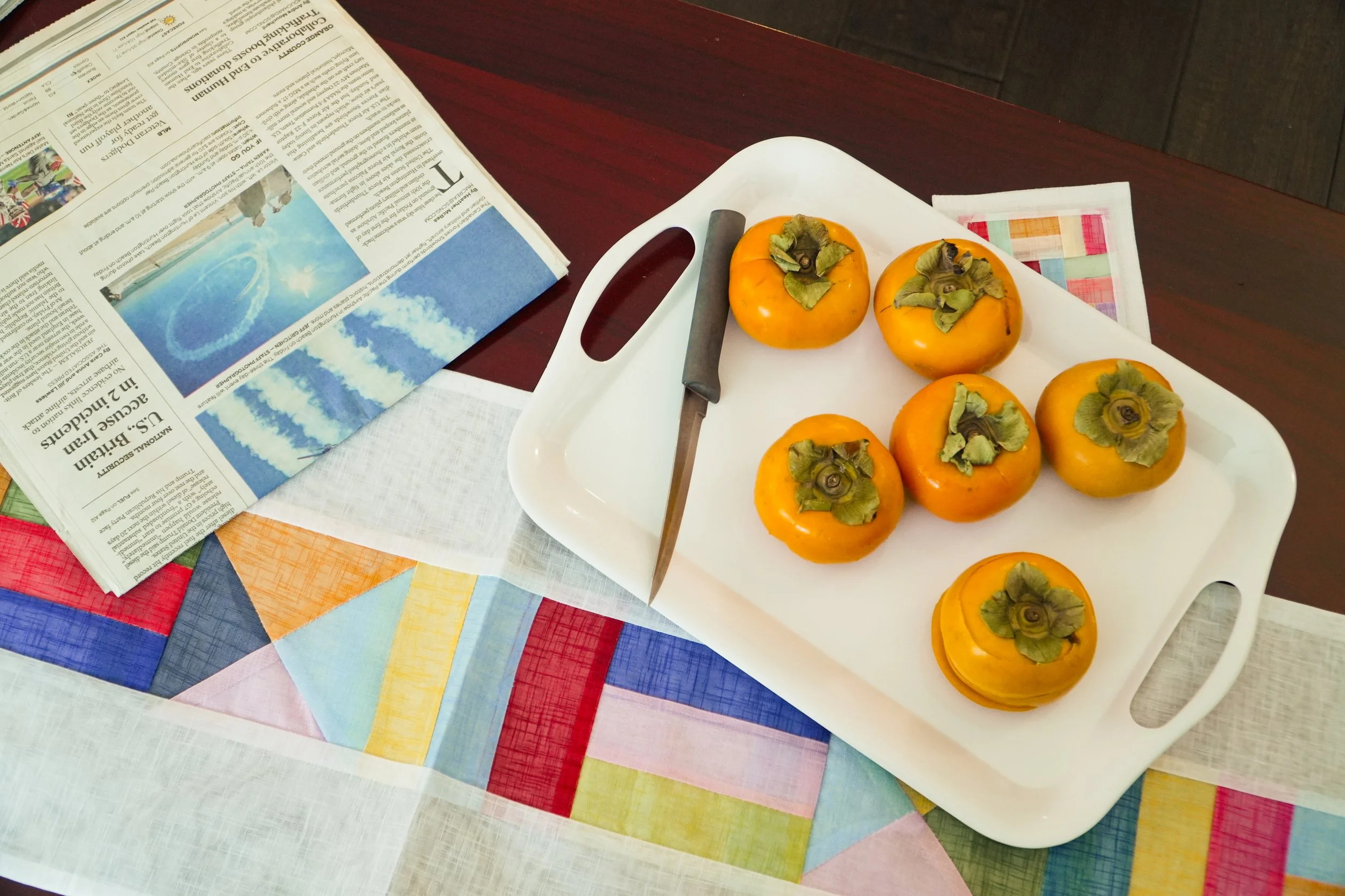 Ripe persimmons on a white serving tray with a knife, newspaper, and colorful quilted tablecloth