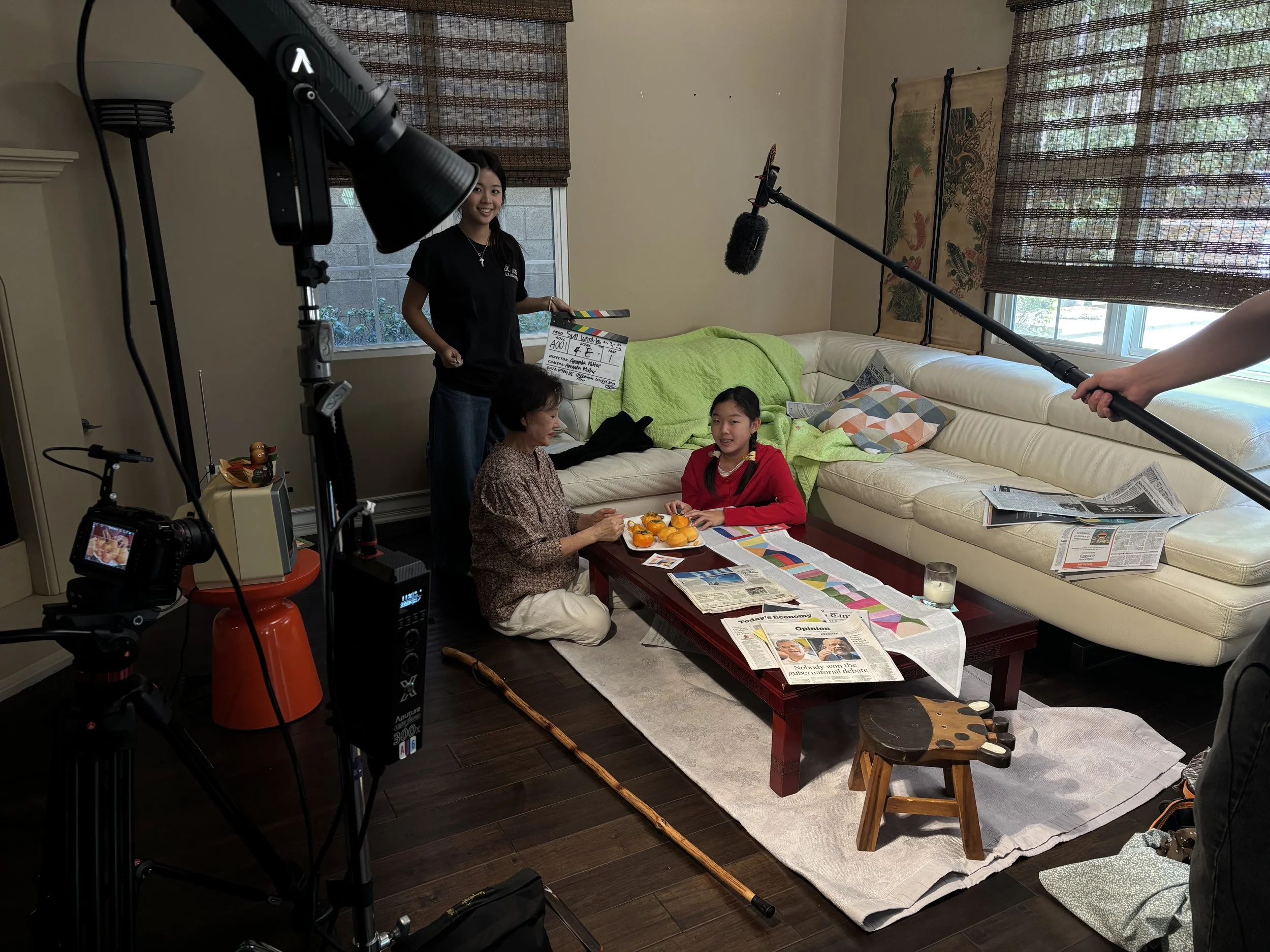 Behind-the-scenes film set in a living room with a camera, boom microphone, lighting equipment, clapperboard, and three people filming a family scene.