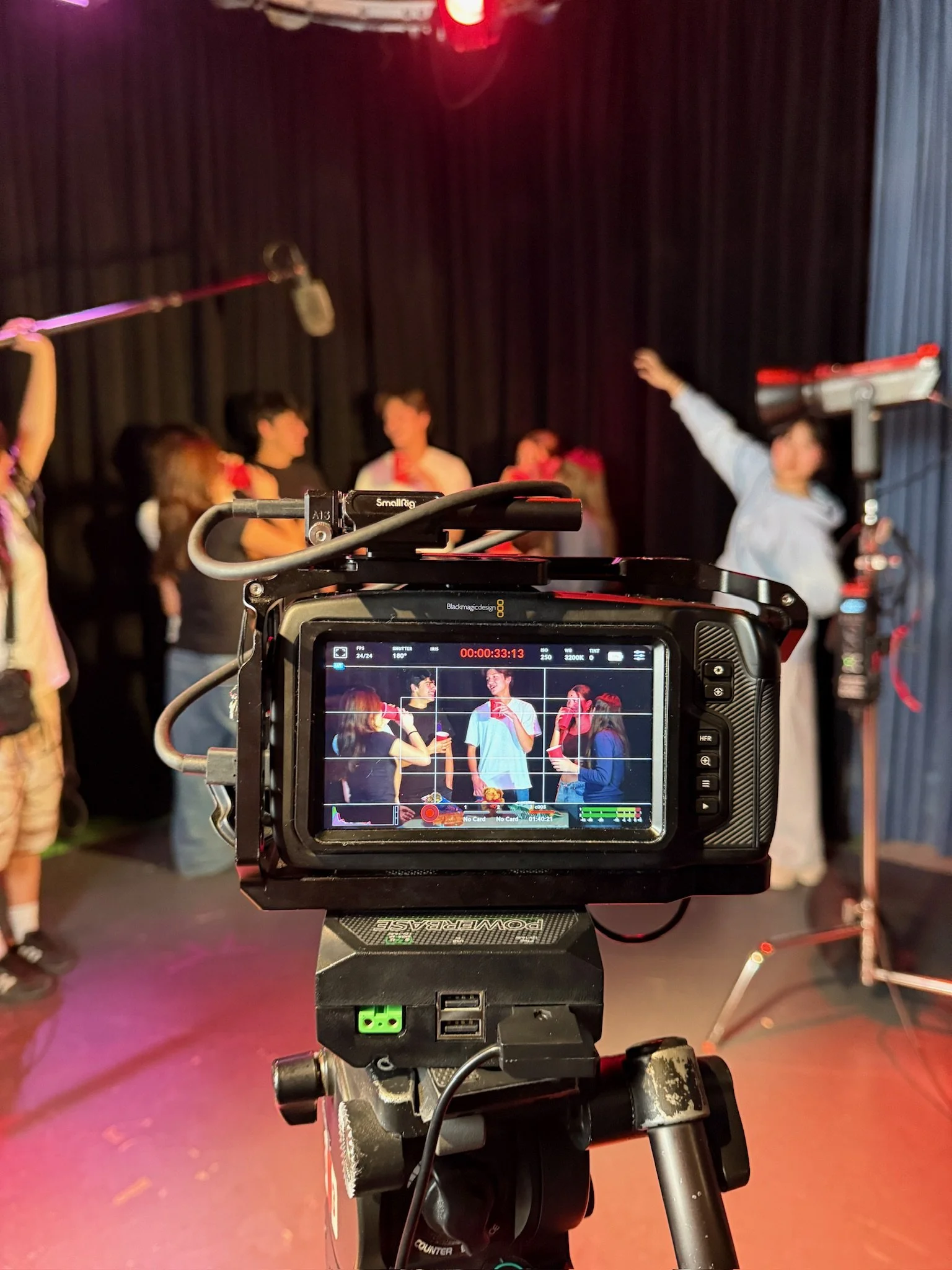 Professional cinema camera filming people on a stage under red lighting, with the camera monitor showing the live scene.