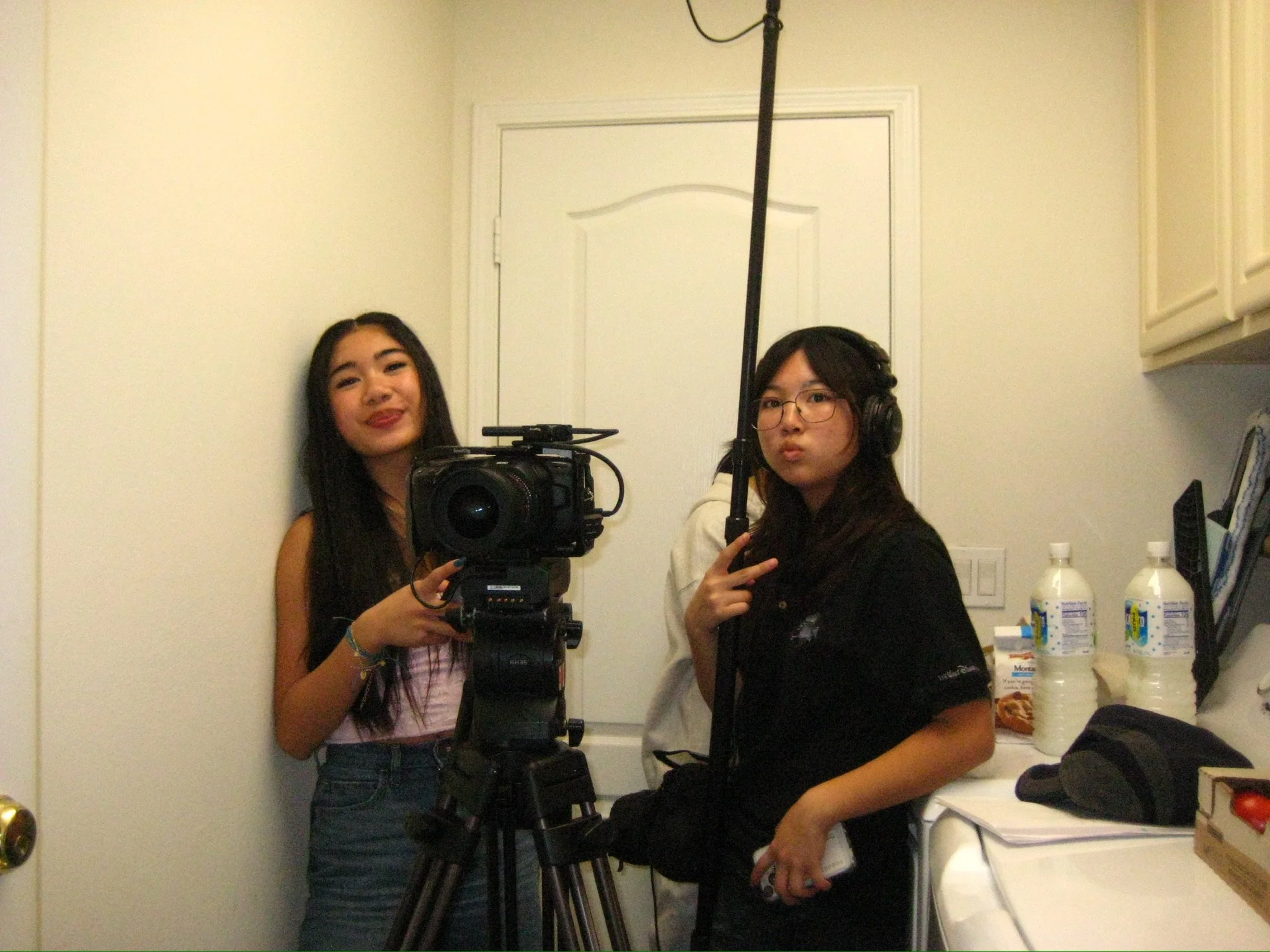 Two women pose indoors beside a professional video camera on a tripod; one wears headphones and holds a microphone.