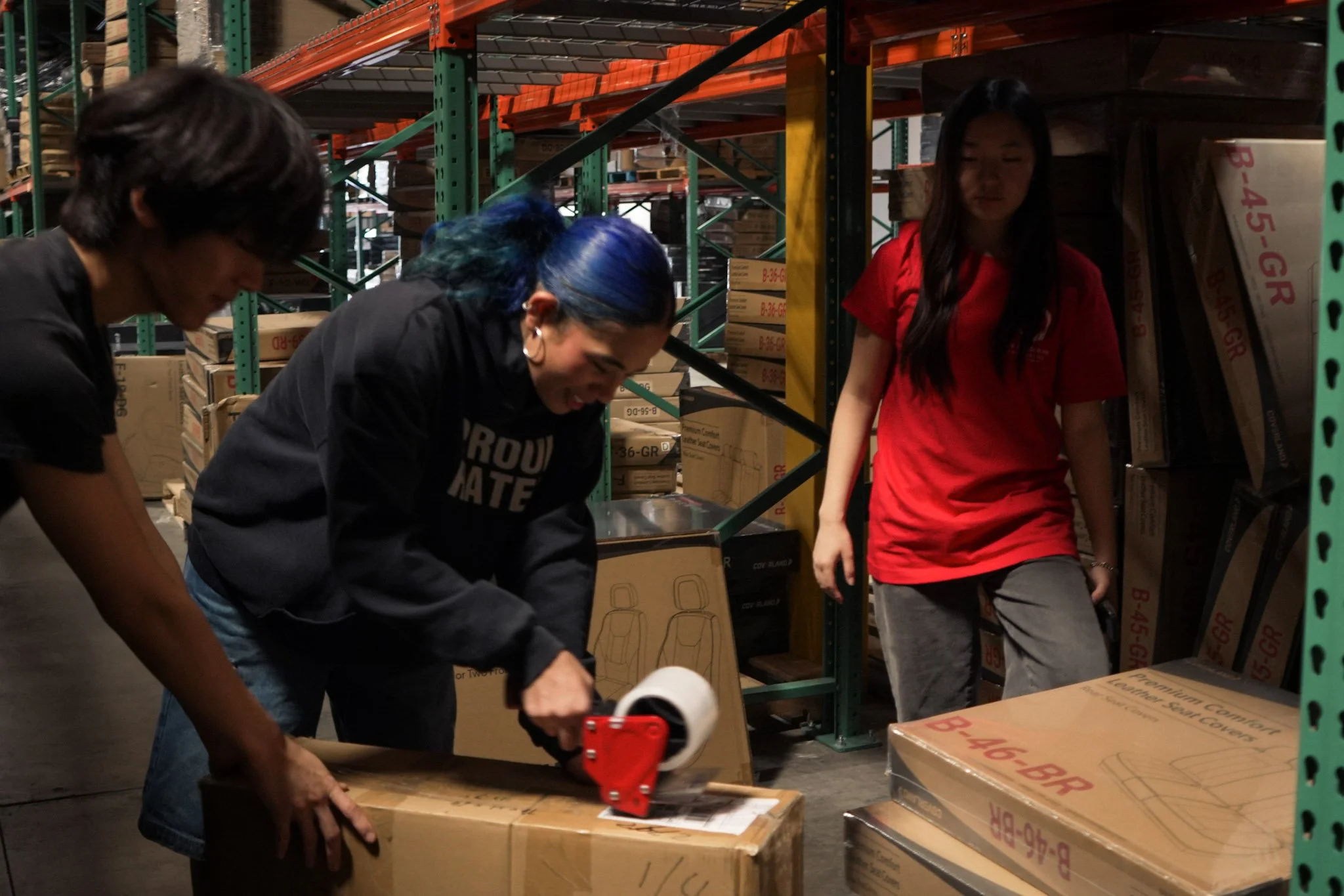 Three workers packing cardboard boxes with a tape dispenser in a warehouse aisle.