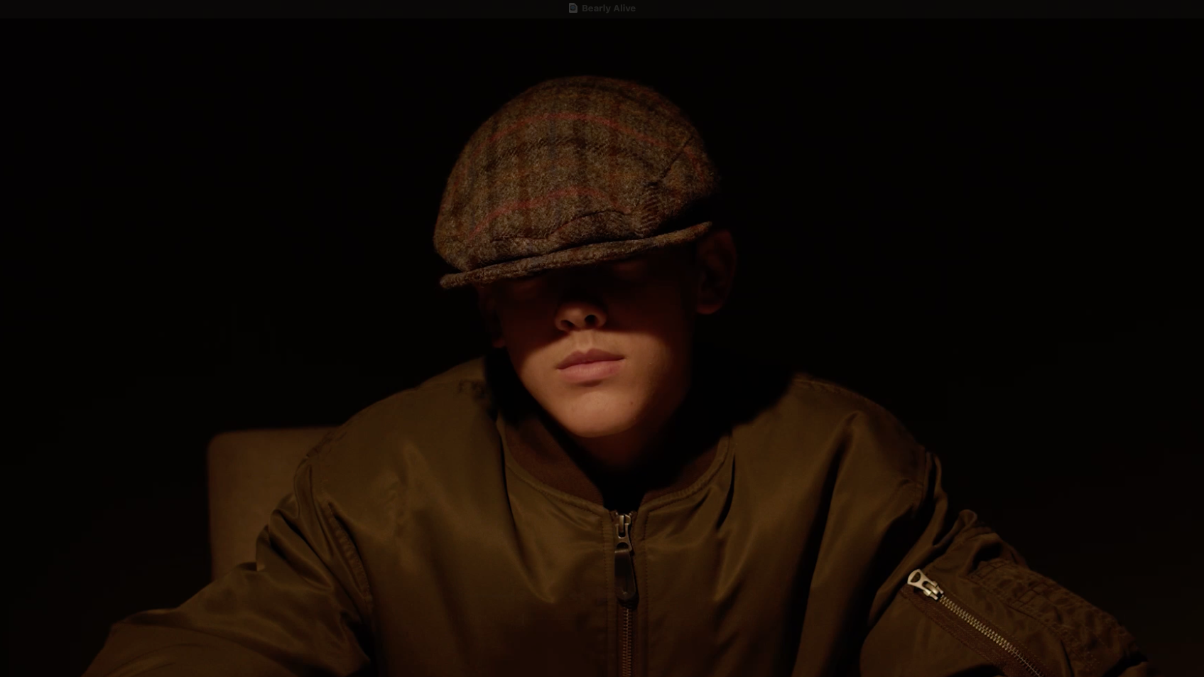 Person wearing a camouflage cap and brown military-style jacket in dim lighting