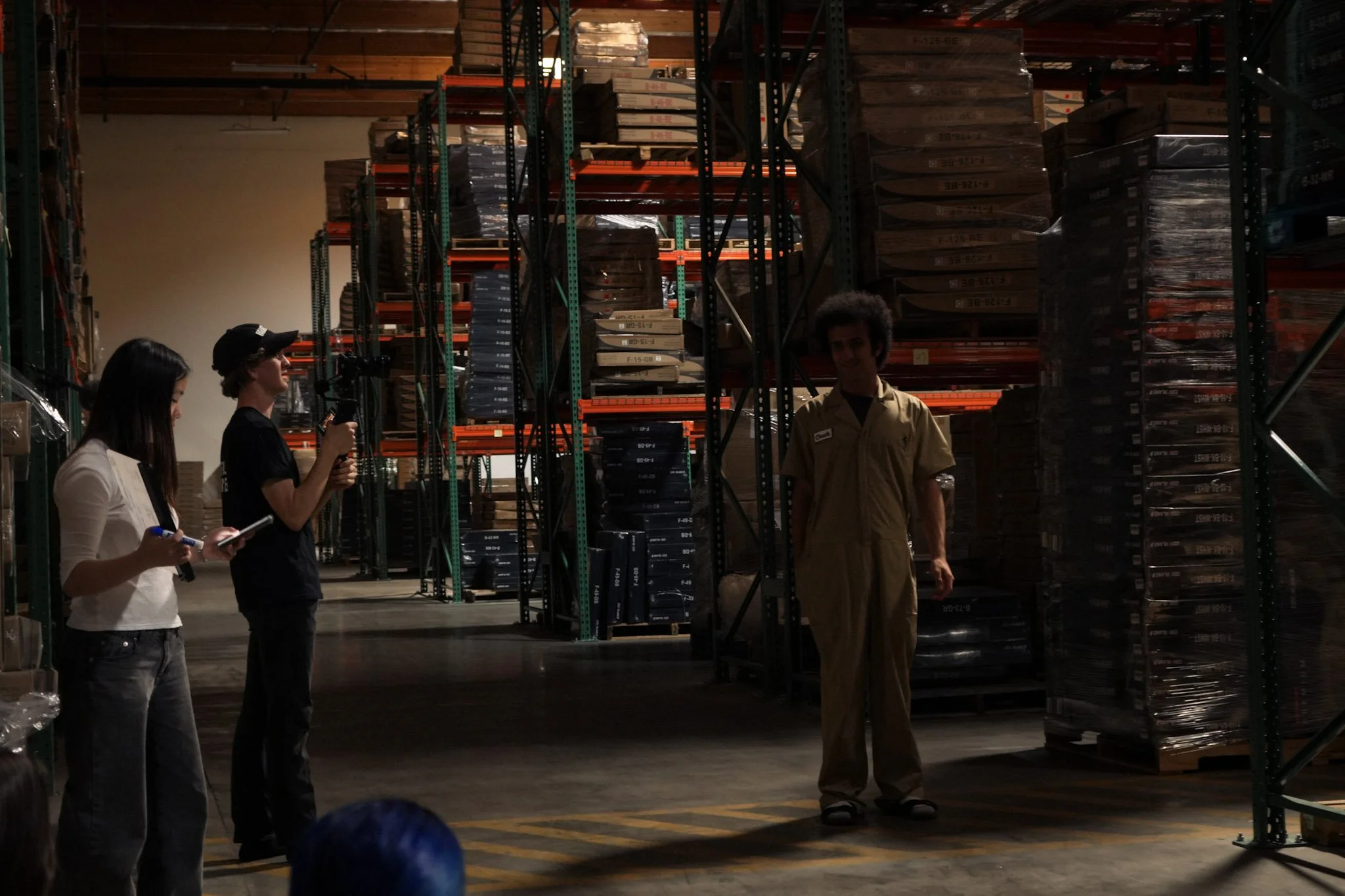 Three people in a warehouse aisle surrounded by tall shelves stacked with packaged materials; one person wears a work uniform while another holds a camera.