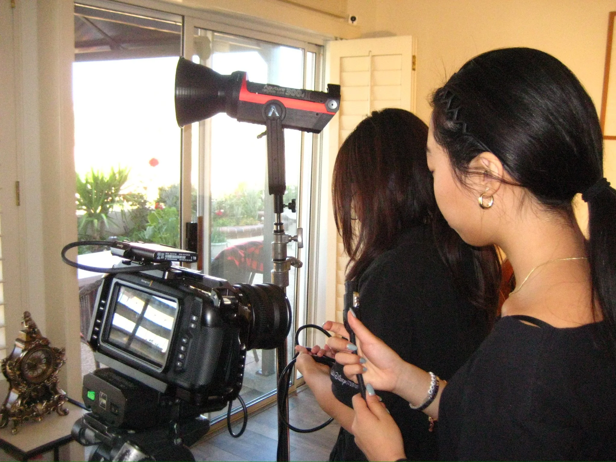 Two women operate a professional video camera with a mounted teleprompter and studio light indoors.