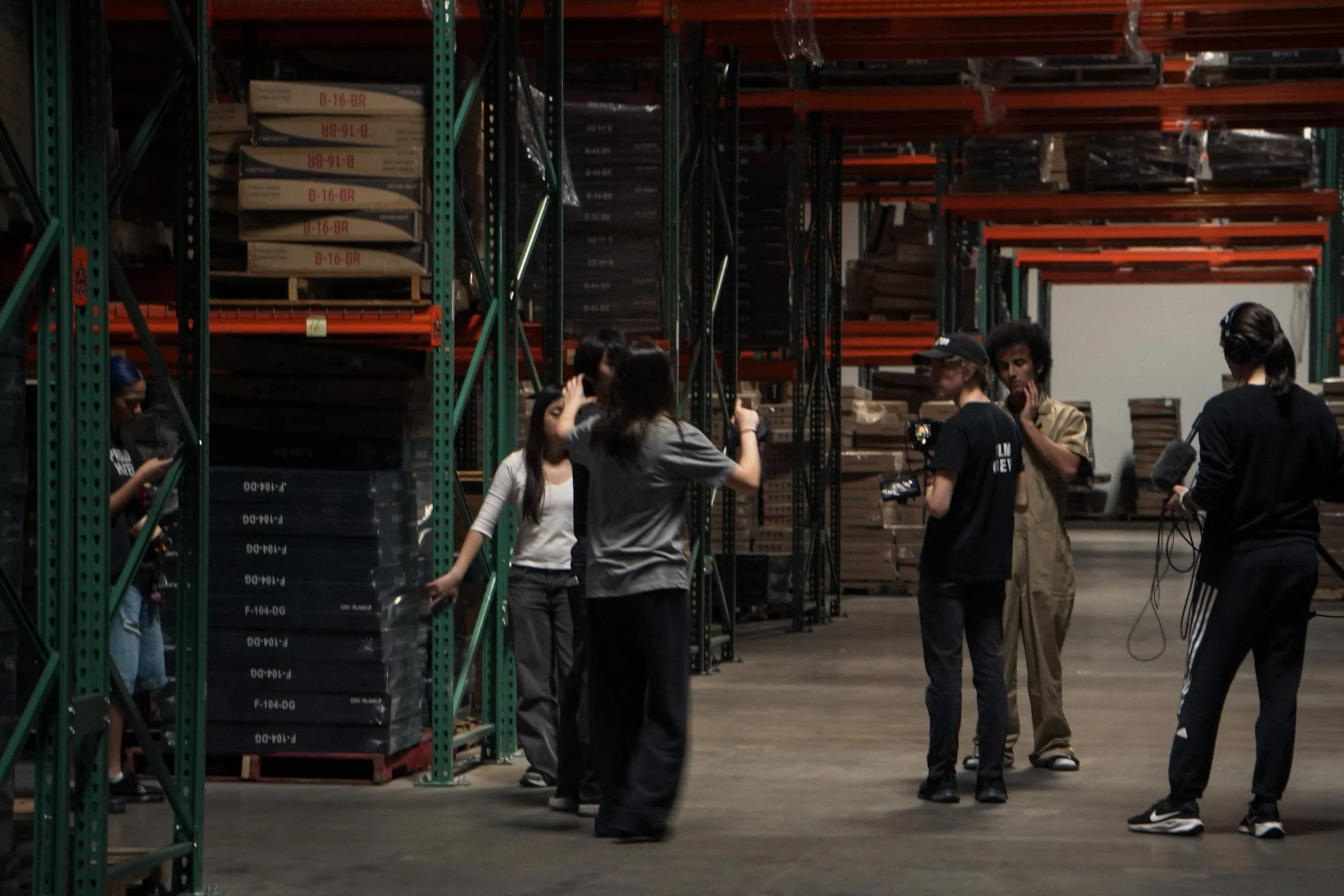Film crew and actors rehearsing in a warehouse with industrial storage racks and boxes