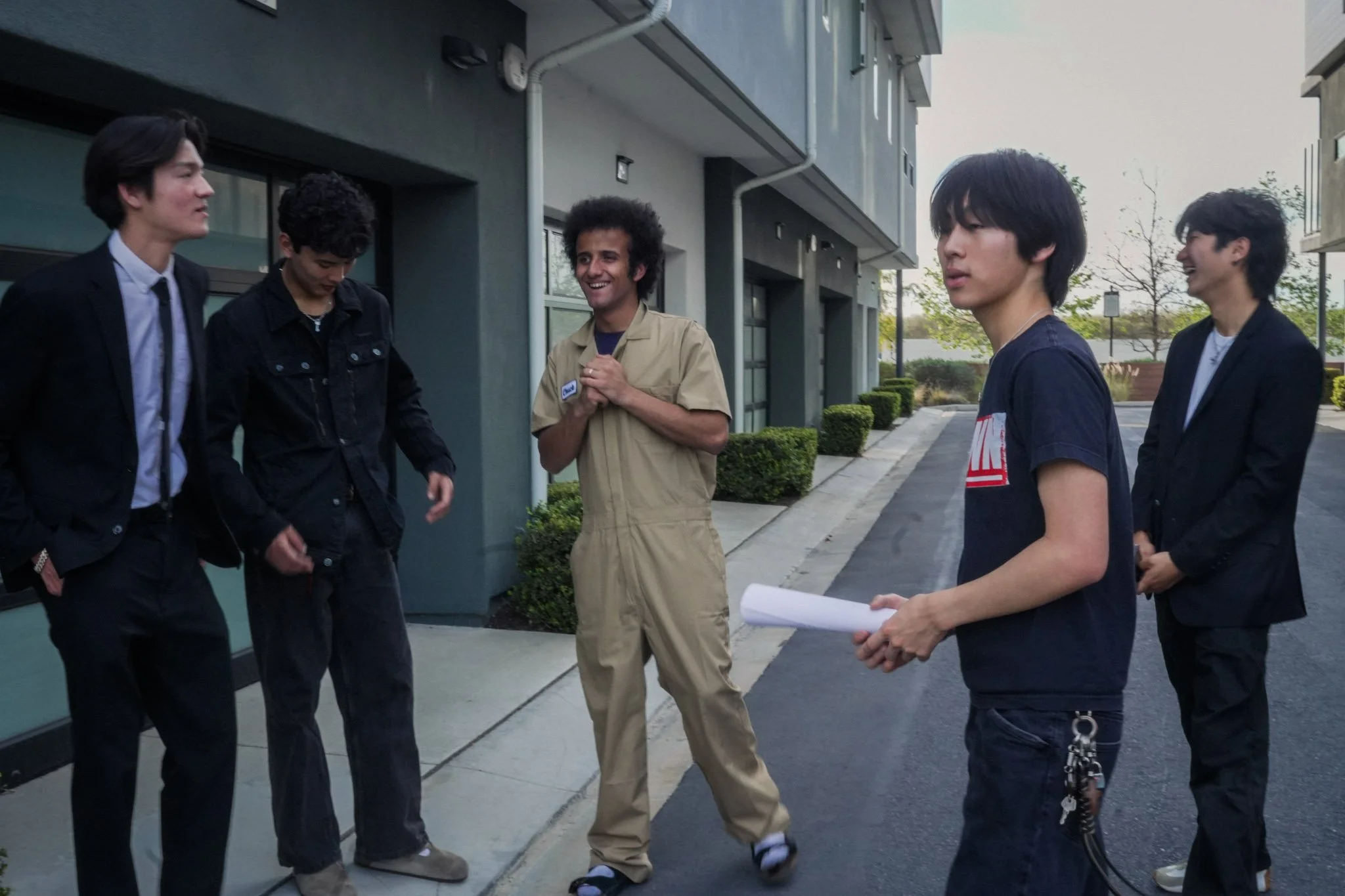 Five young men talking outside a modern building, with one holding rolled papers.