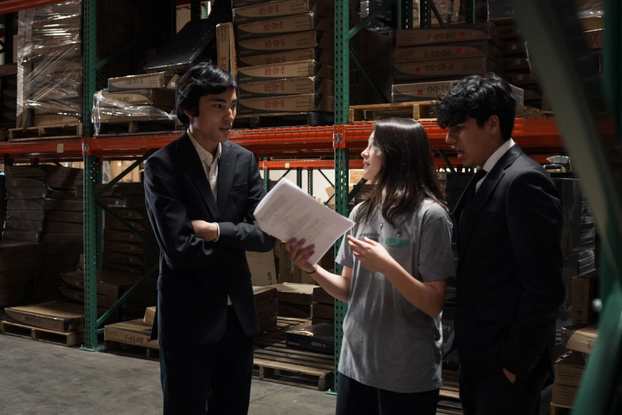 Three coworkers discuss paperwork in a warehouse aisle surrounded by stacked boxes and industrial shelving.