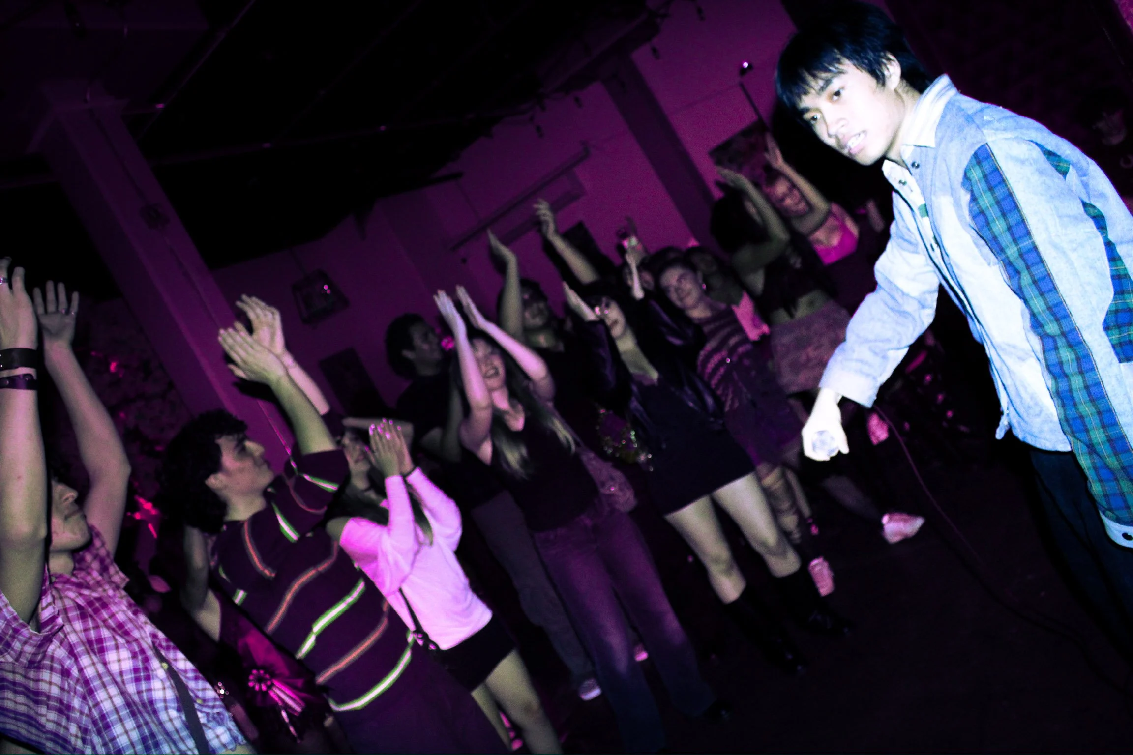 Crowd dancing with raised hands under purple nightclub lighting as a man in a plaid shirt leans toward the camera.
