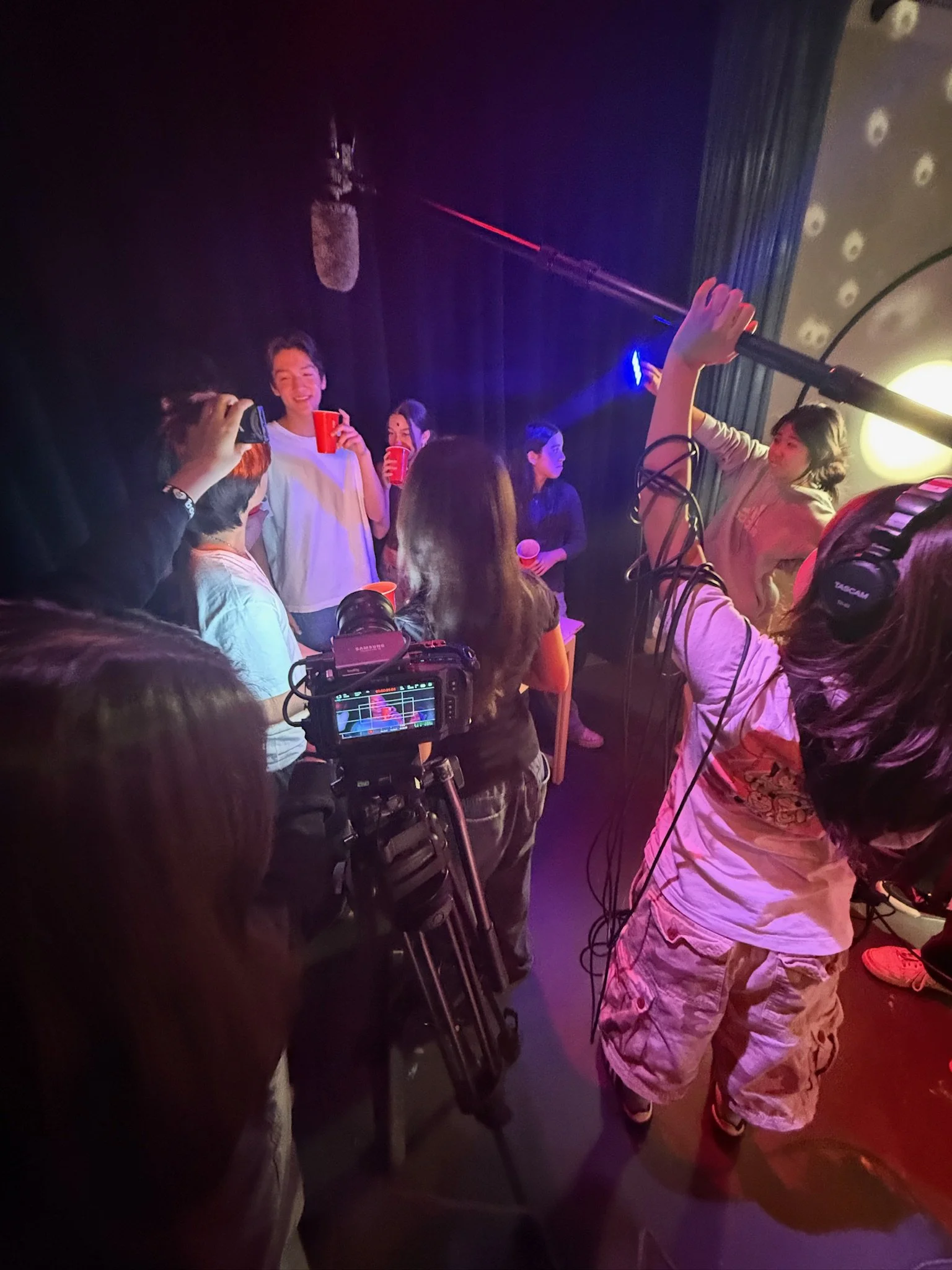 Film crew recording people at a dimly lit party with a boom microphone and professional video camera.