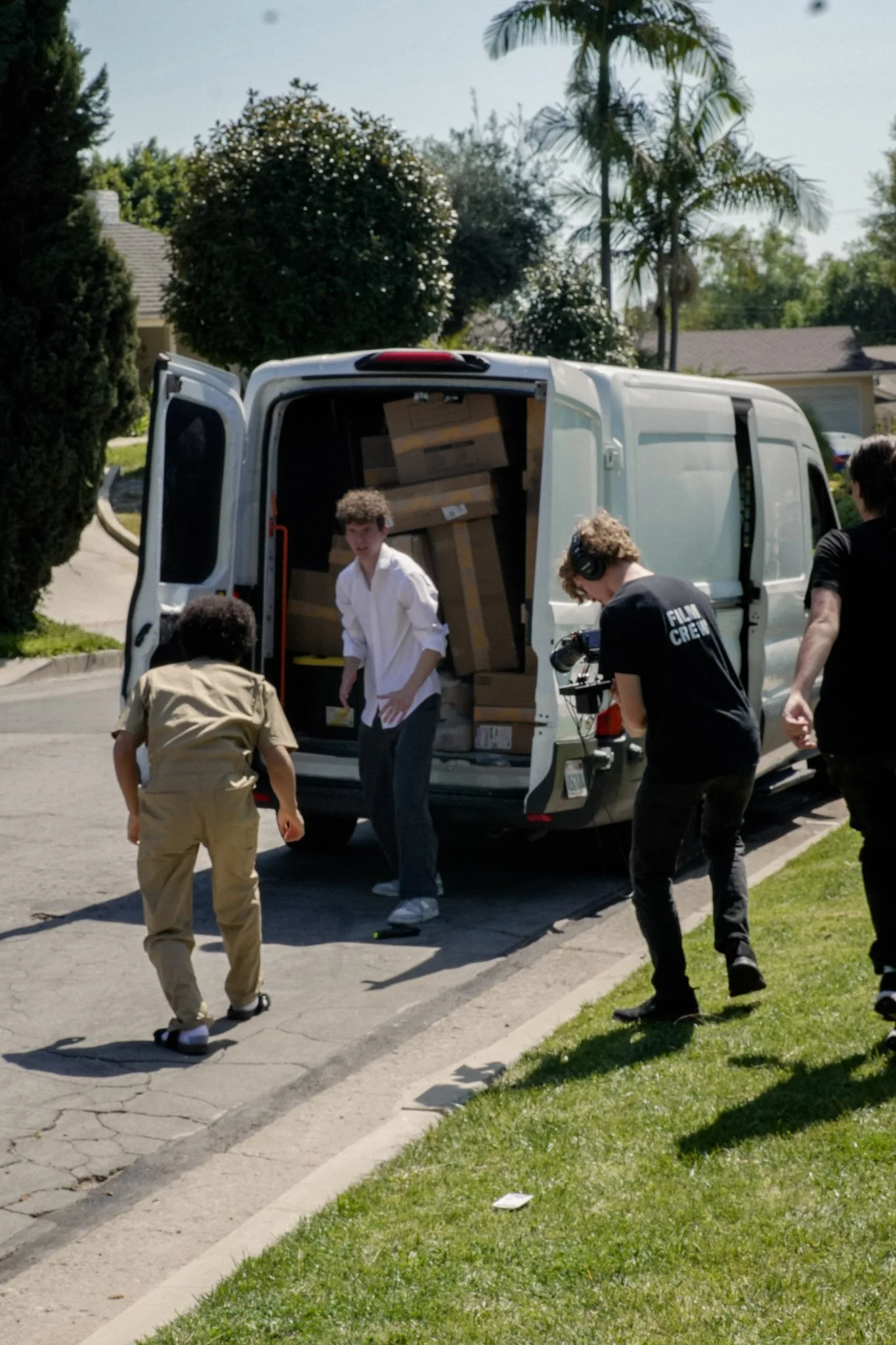 Film crew unloading cardboard boxes from a white cargo van on a suburban street.