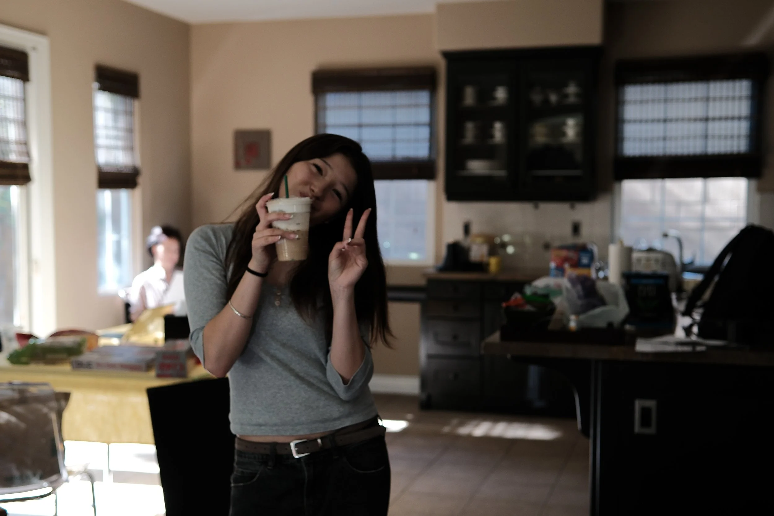 Woman drinking a blended beverage and making a peace sign in a sunlit kitchen