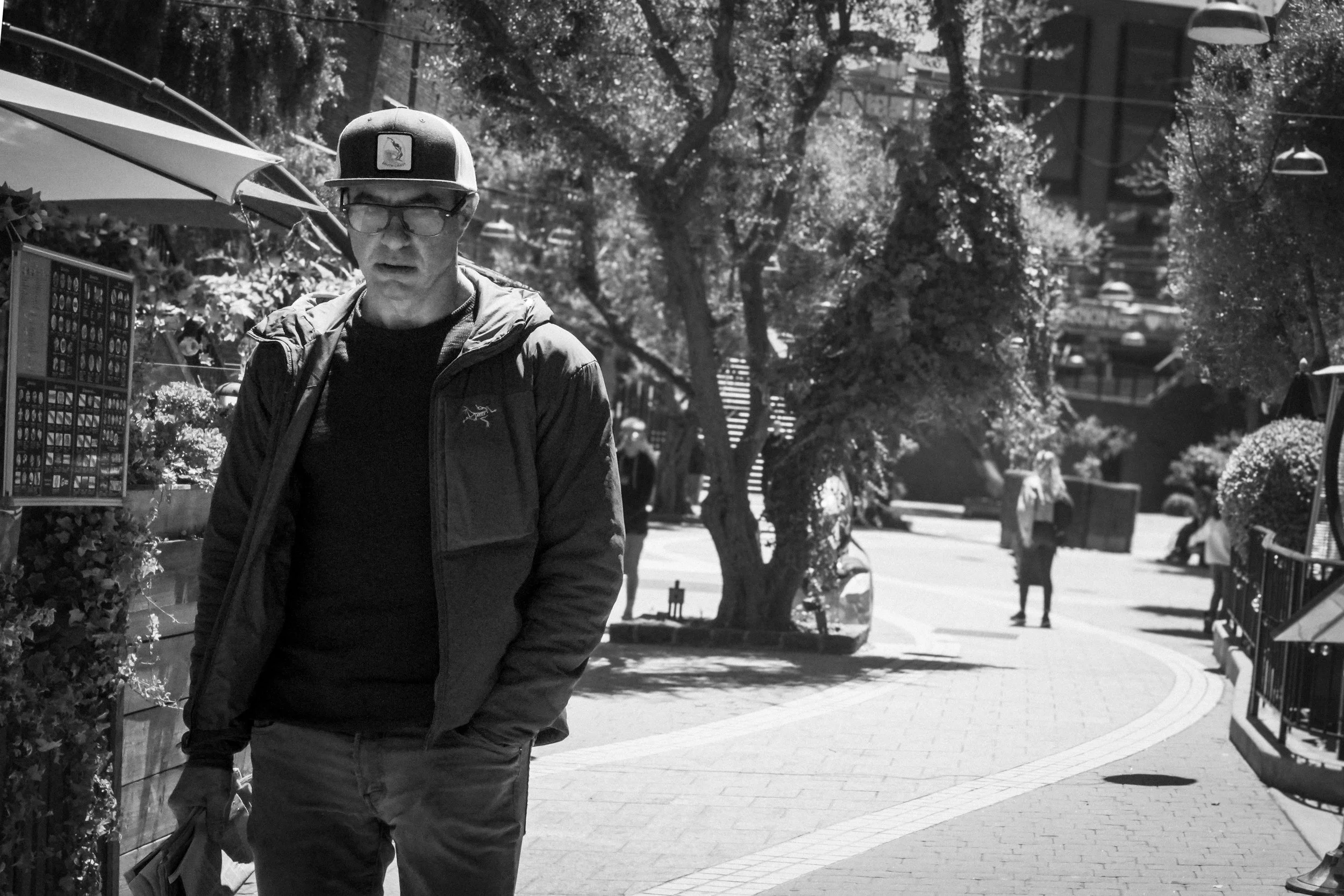 Man wearing glasses, a baseball cap, and jacket standing on a tree-lined outdoor shopping walkway with umbrellas and pedestrians.