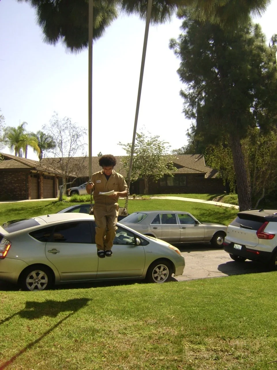 Man in a uniform standing on a parked green Toyota Prius, holding two long poles in a suburban neighborhood