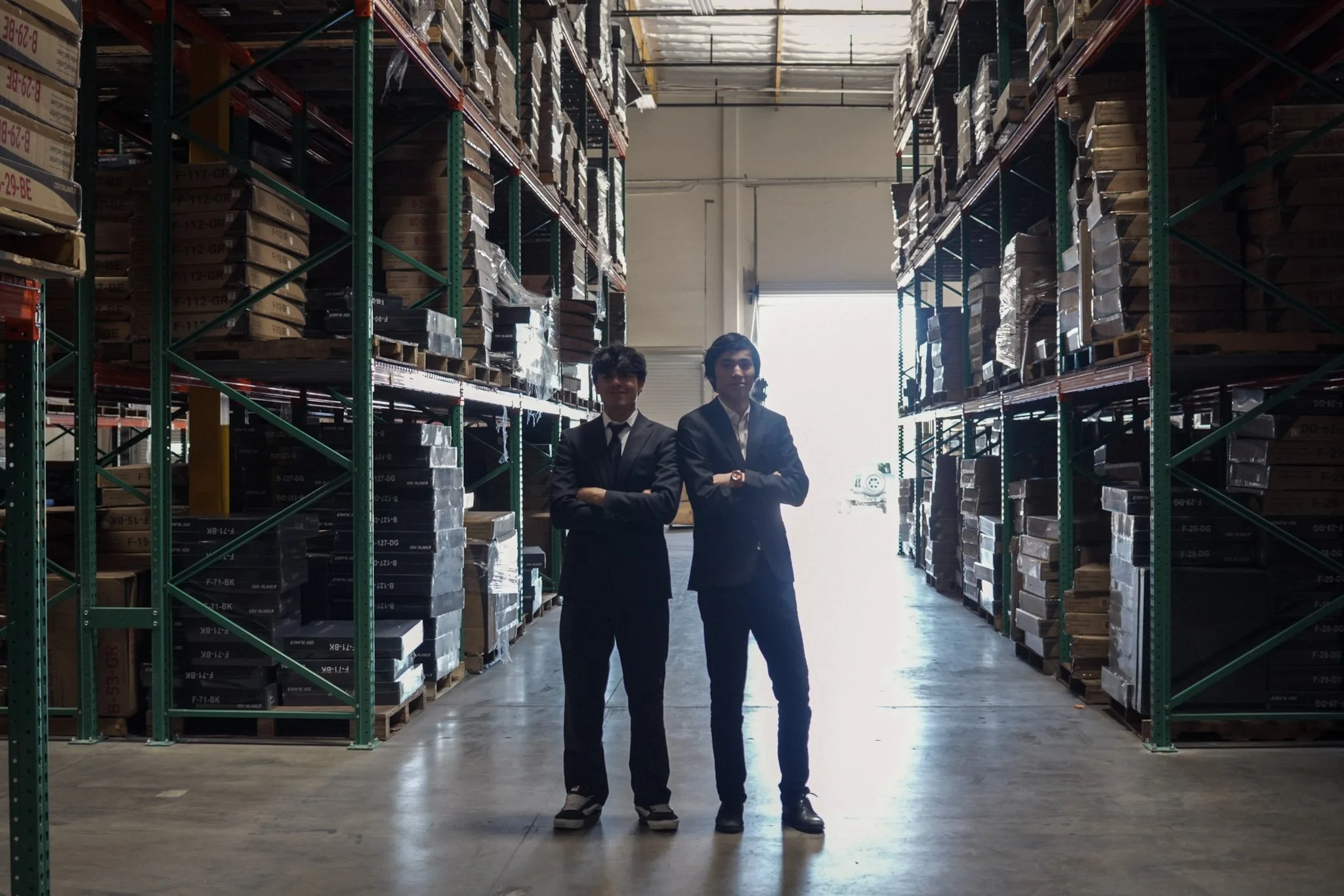 Two men in suits standing with folded arms in a warehouse aisle surrounded by tall racks of boxed inventory.