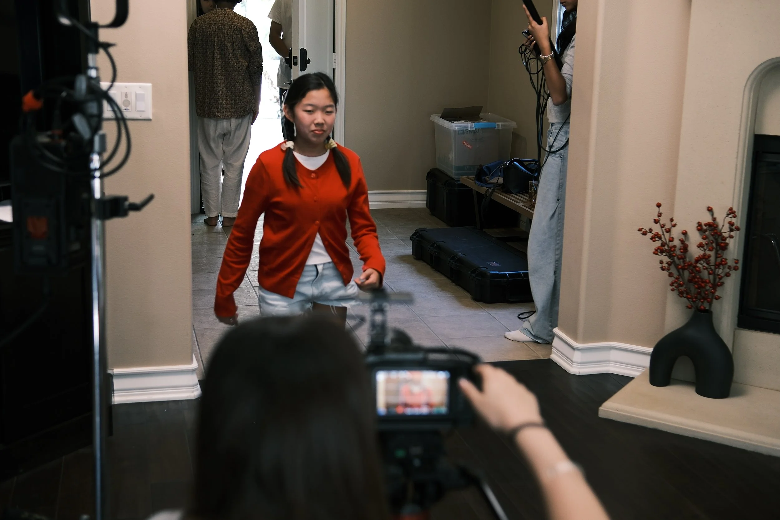 Young woman in a red cardigan filmed indoors by a camera crew, with production equipment visible in the foreground.