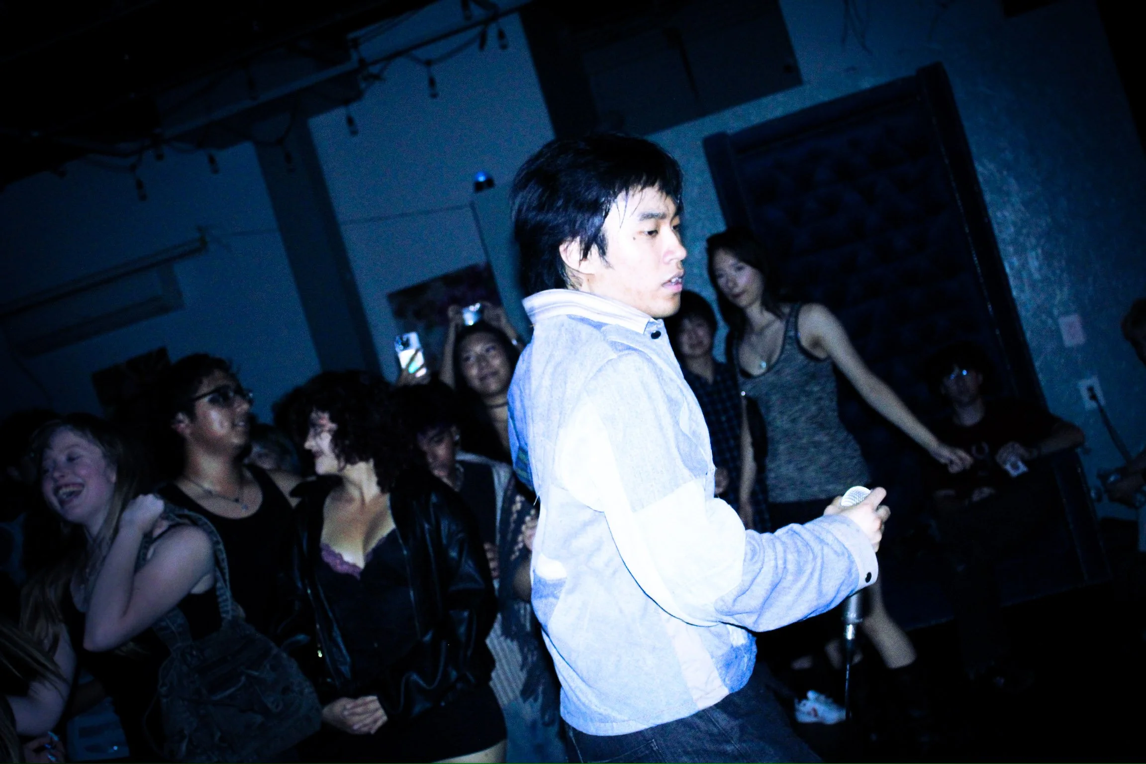 Singer holding a microphone performs among an enthusiastic crowd at a dimly lit concert.