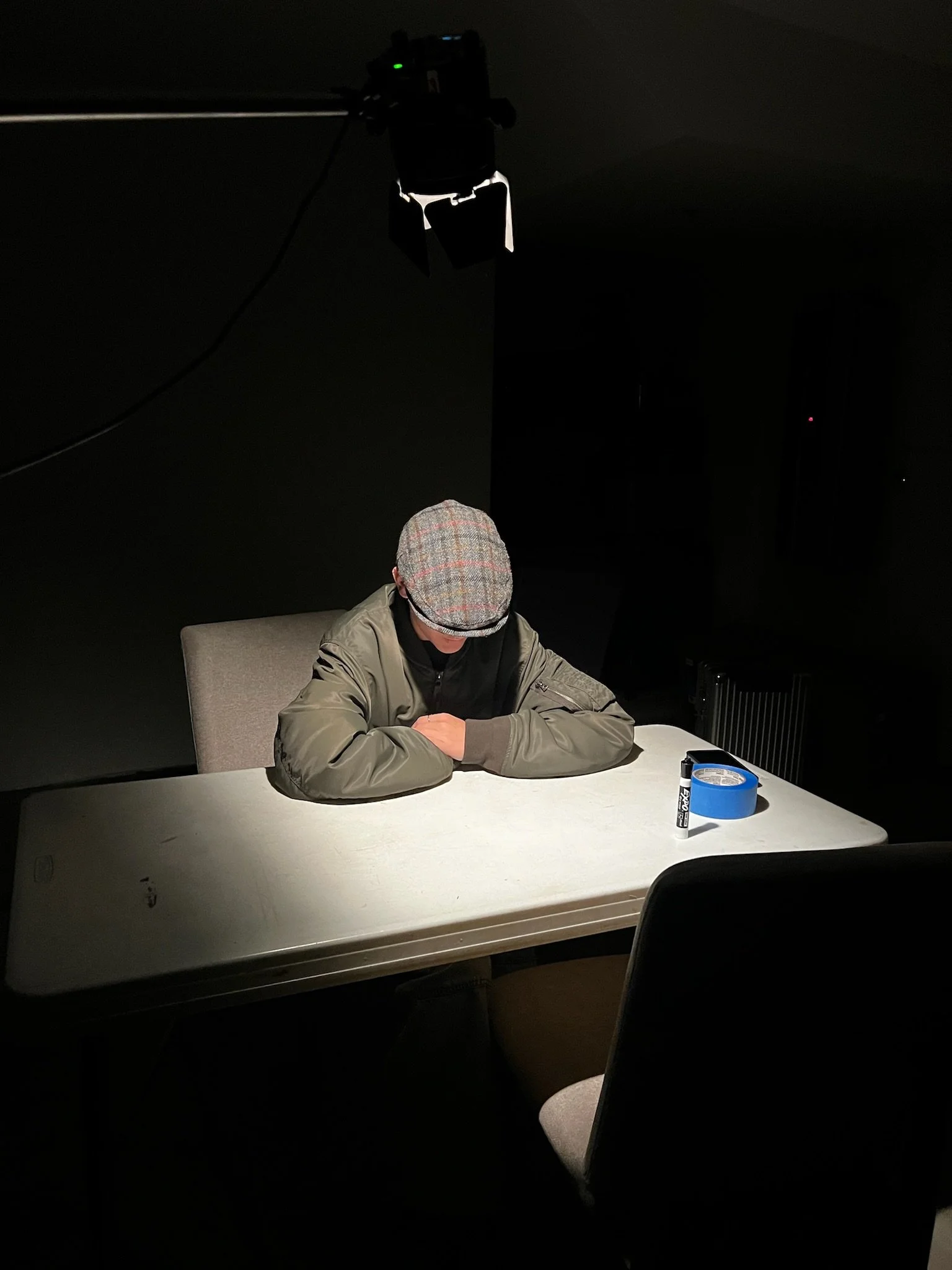 Person in a flat cap and jacket sitting at a table in a dark room, with blue tape and a marker nearby