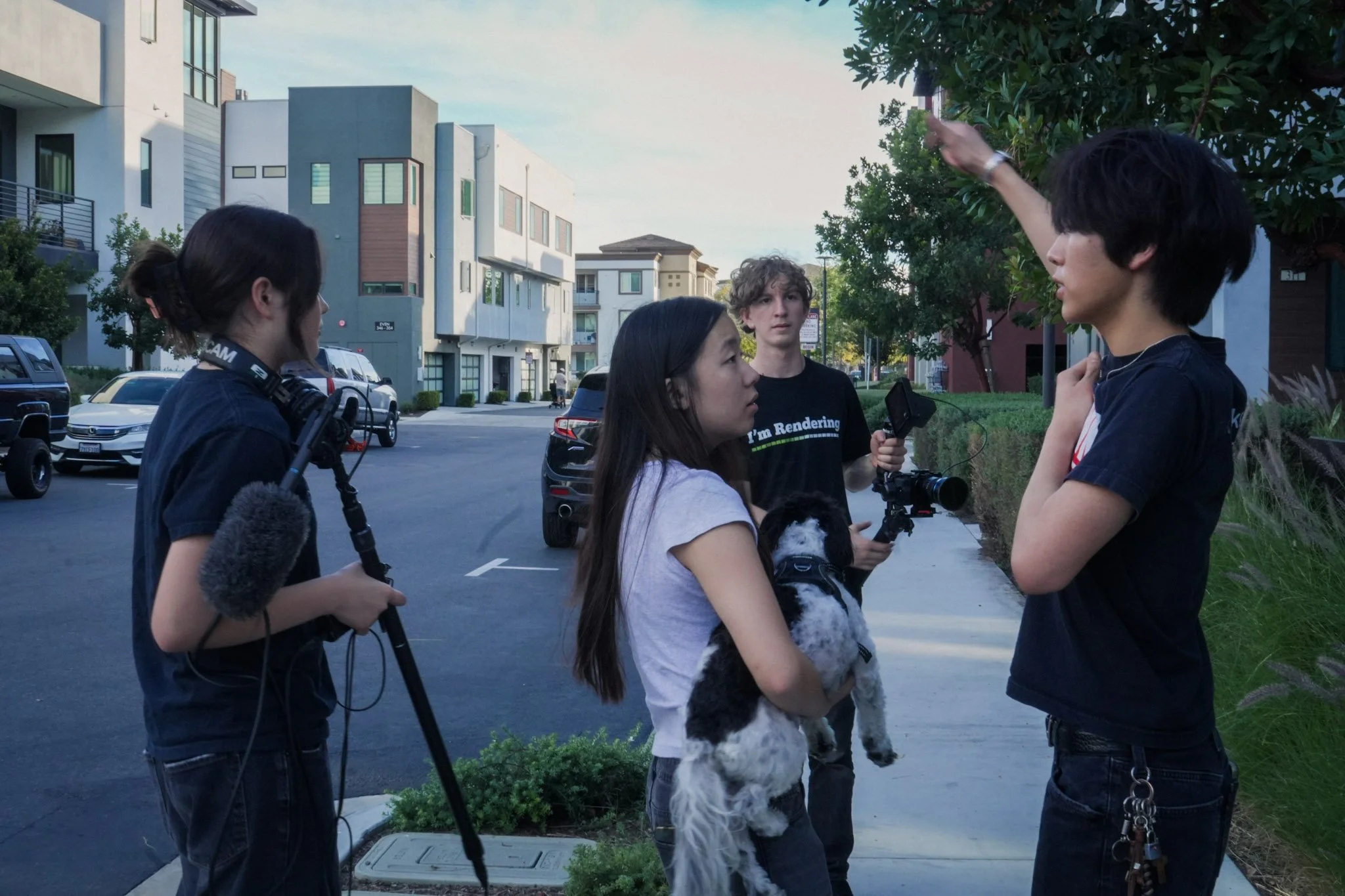 Film crew interviewing a man on a residential street while a woman holds a small black-and-white dog.