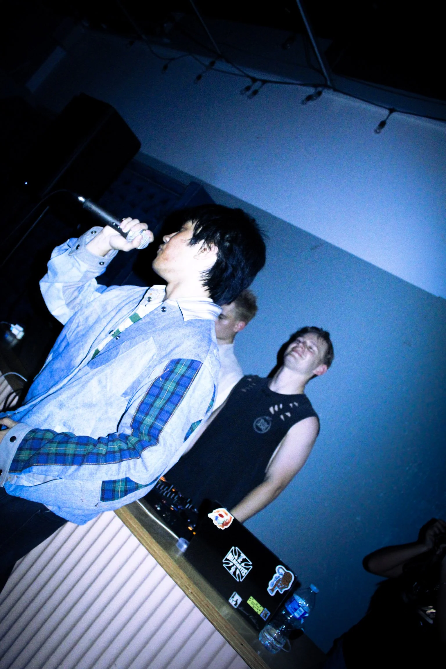 Singer performing into a microphone onstage beside a guitarist with a sticker-covered instrument case.
