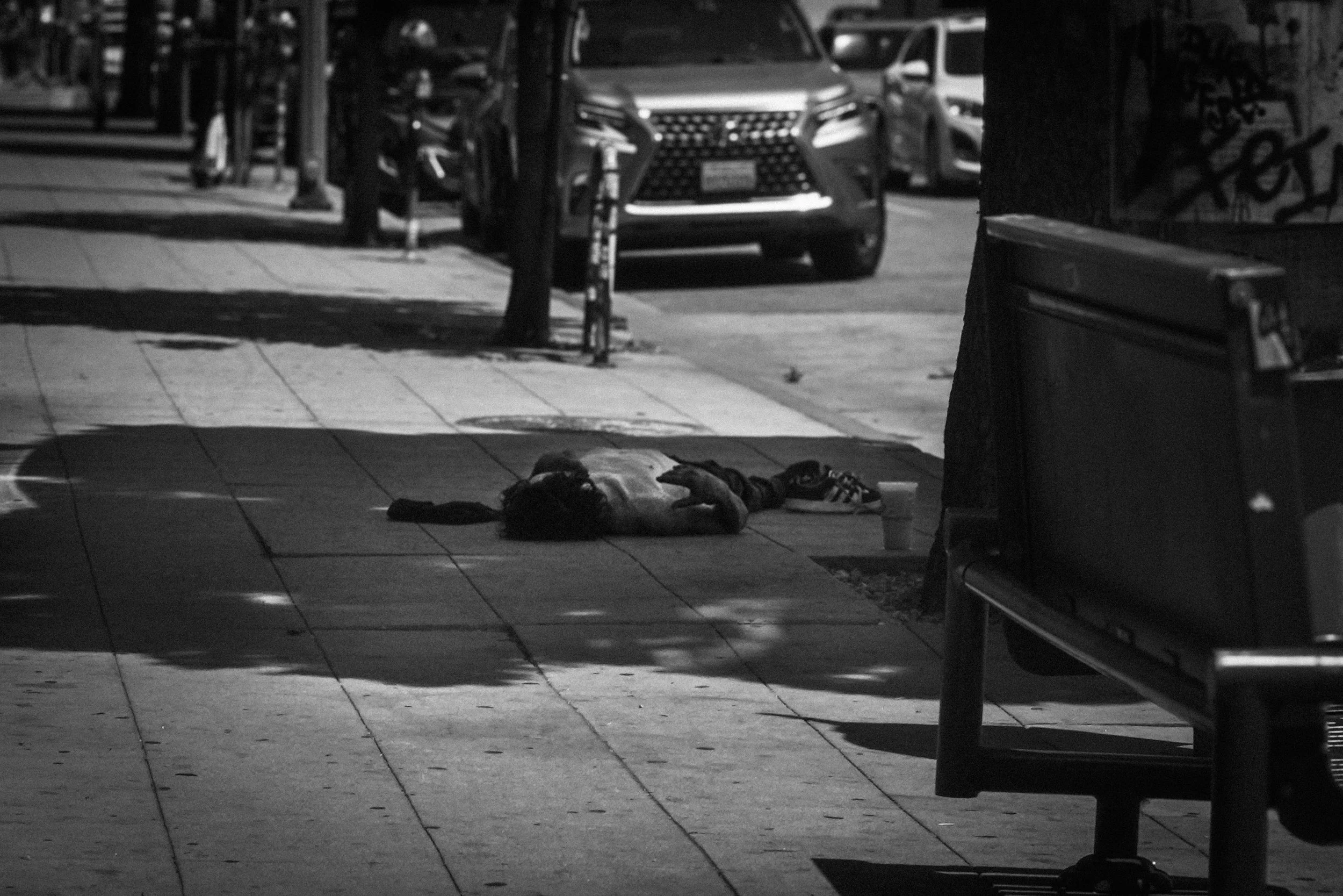 Black-and-white street scene with a person lying on the sidewalk, a parked car, and a bench.