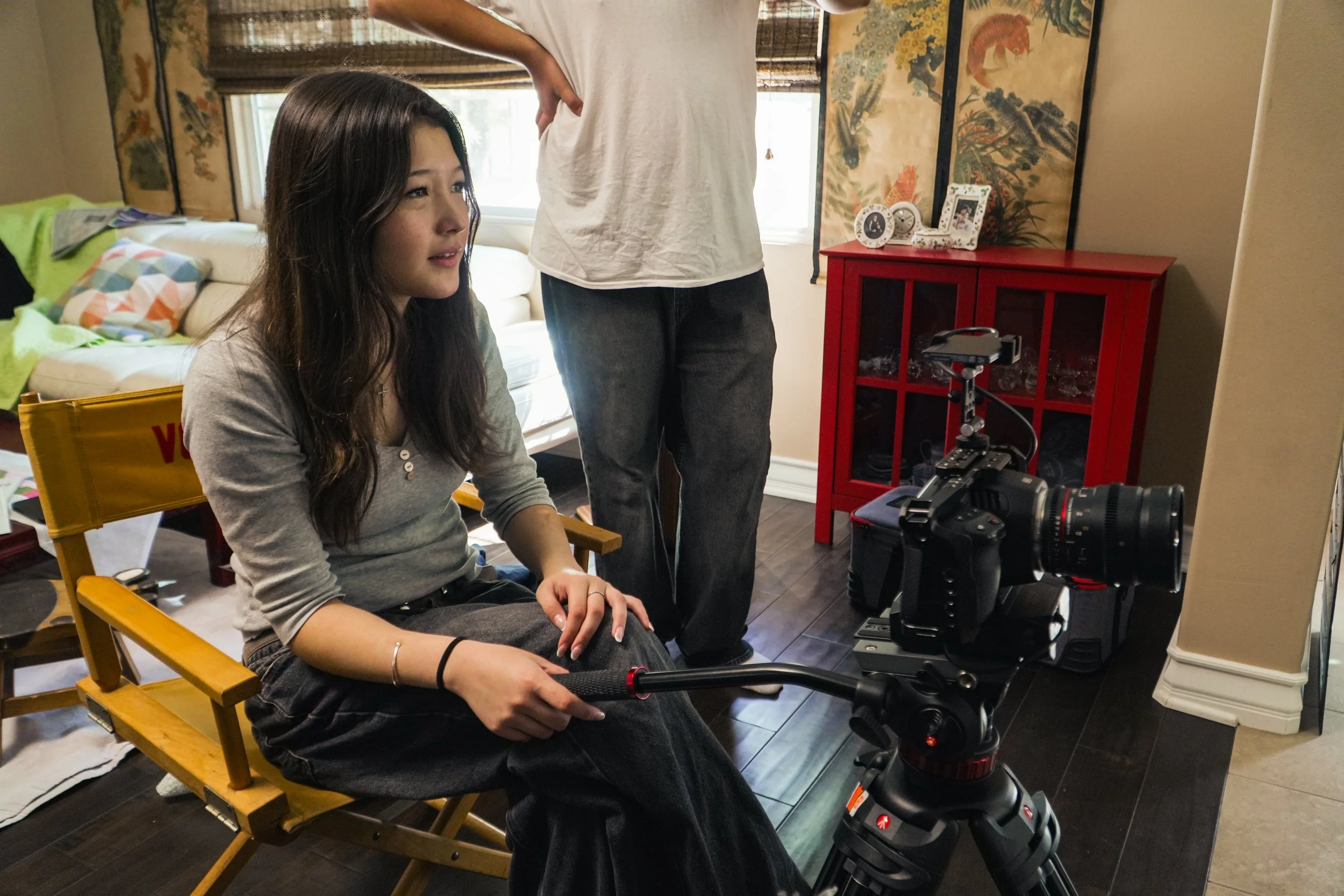 Woman seated in a director’s chair beside a professional camera rig, with a person standing behind her in a living room.