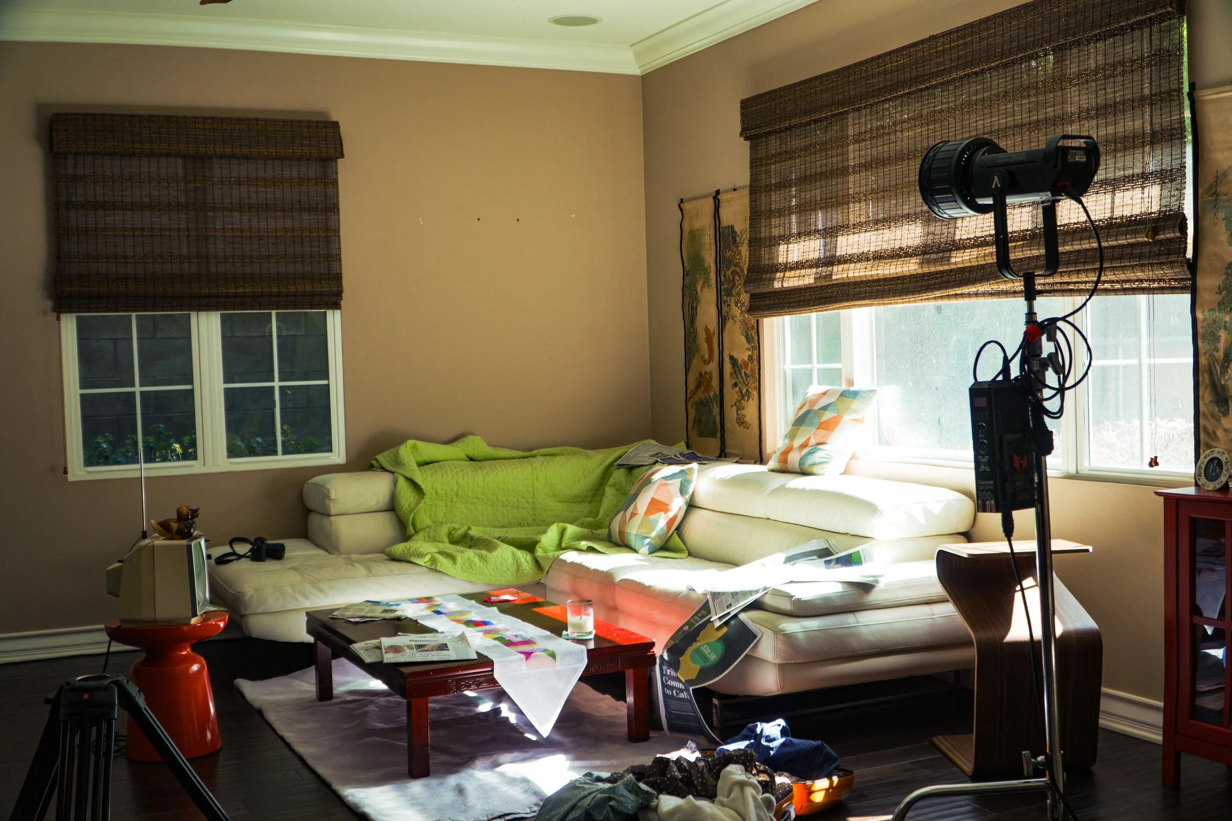 Sunlit living room with a white sectional sofa, green throw blanket, coffee table, large windows, and professional video lighting equipment.