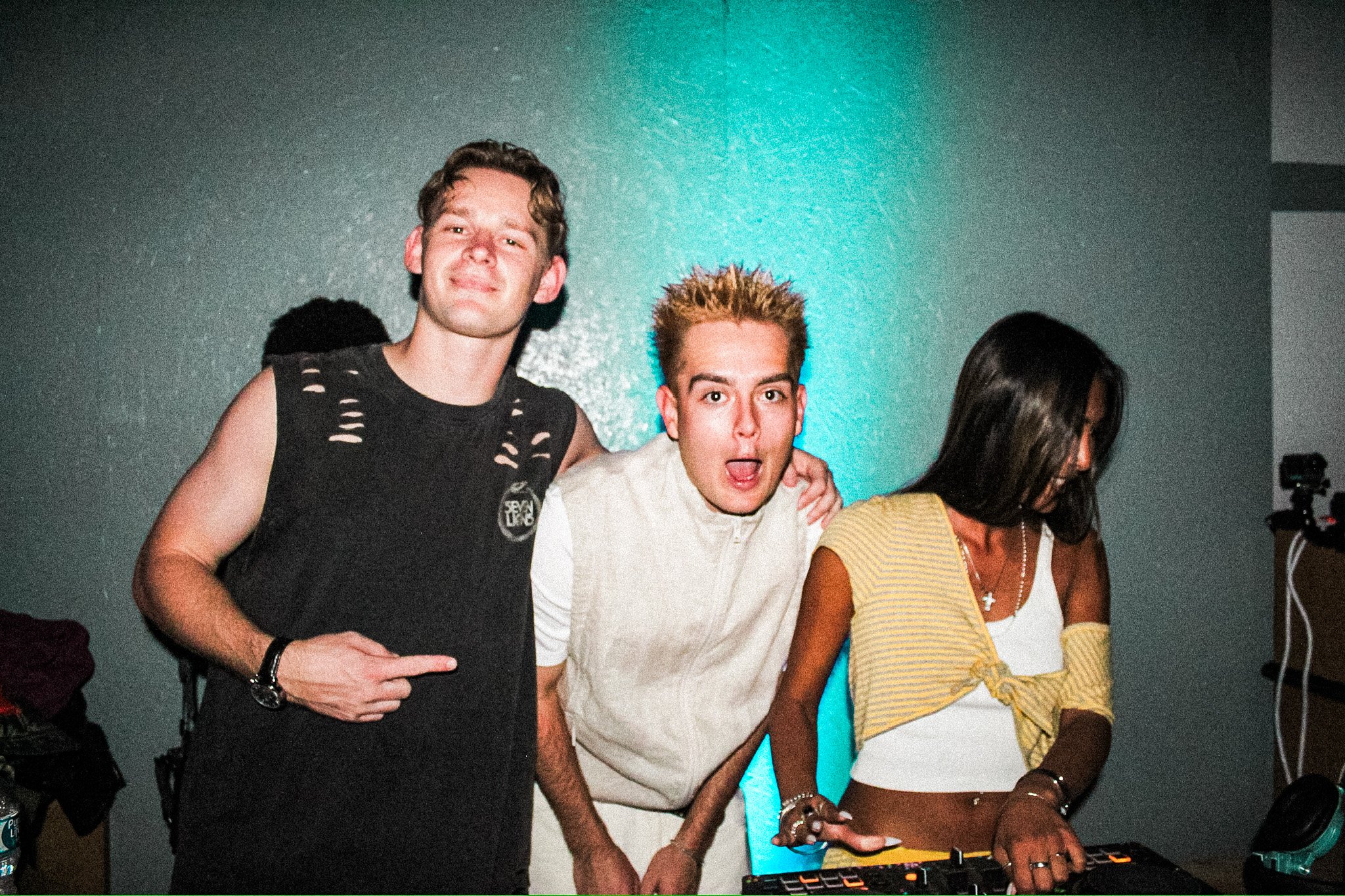 Three people posing indoors near DJ equipment, with turquoise lighting in the background