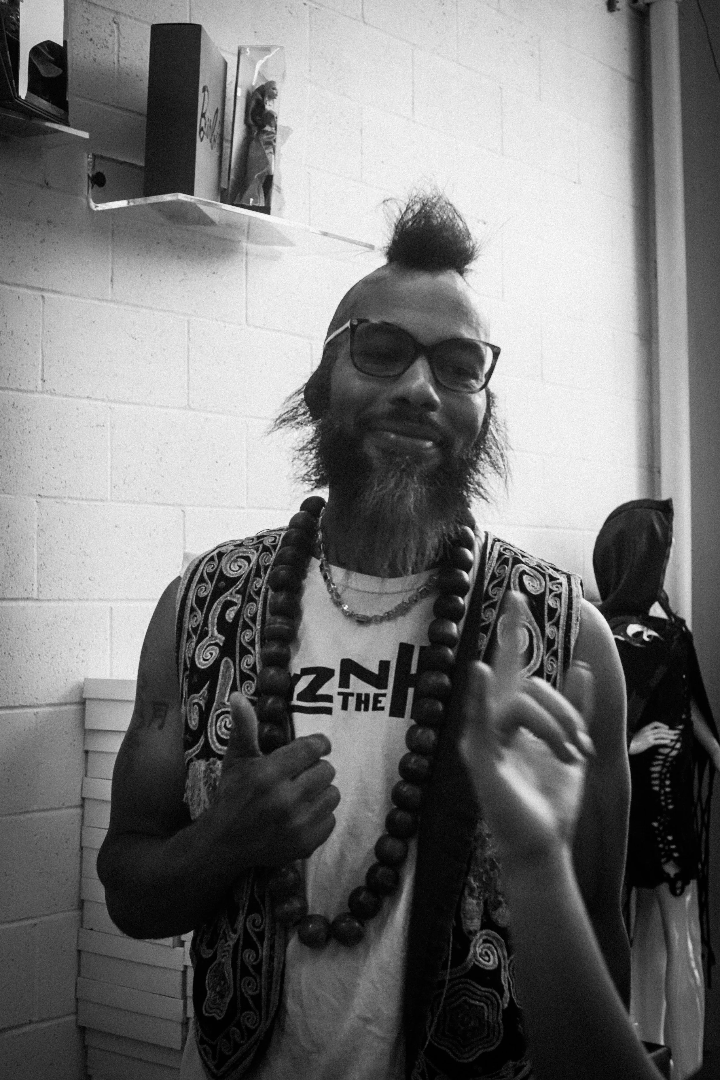 Black-and-white portrait of a bearded man wearing sunglasses, a patterned vest, large beaded necklace, and graphic T-shirt indoors.