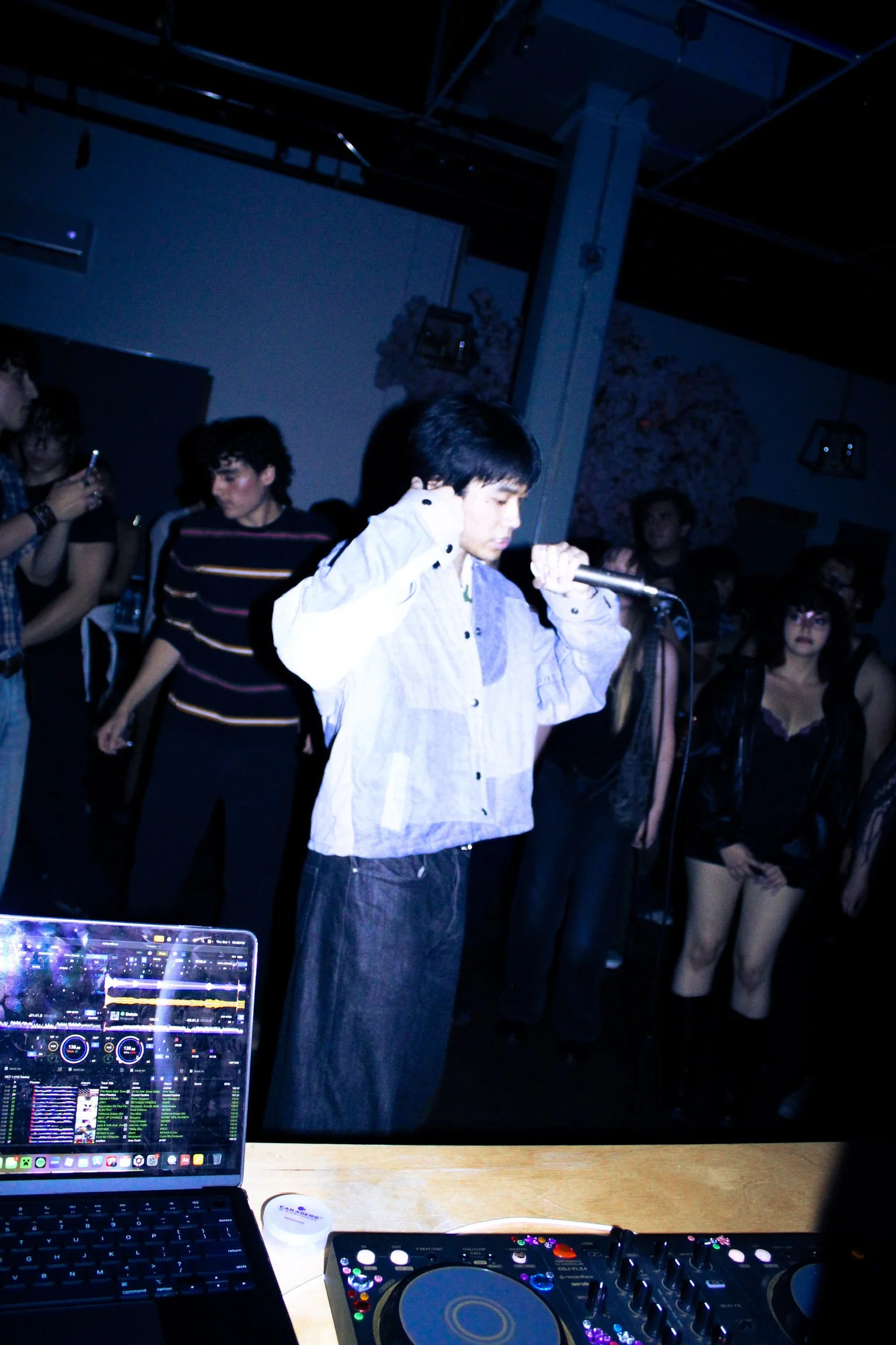 Singer performing into a microphone at a dimly lit nightclub, with dancers, a laptop, and DJ equipment in the foreground.