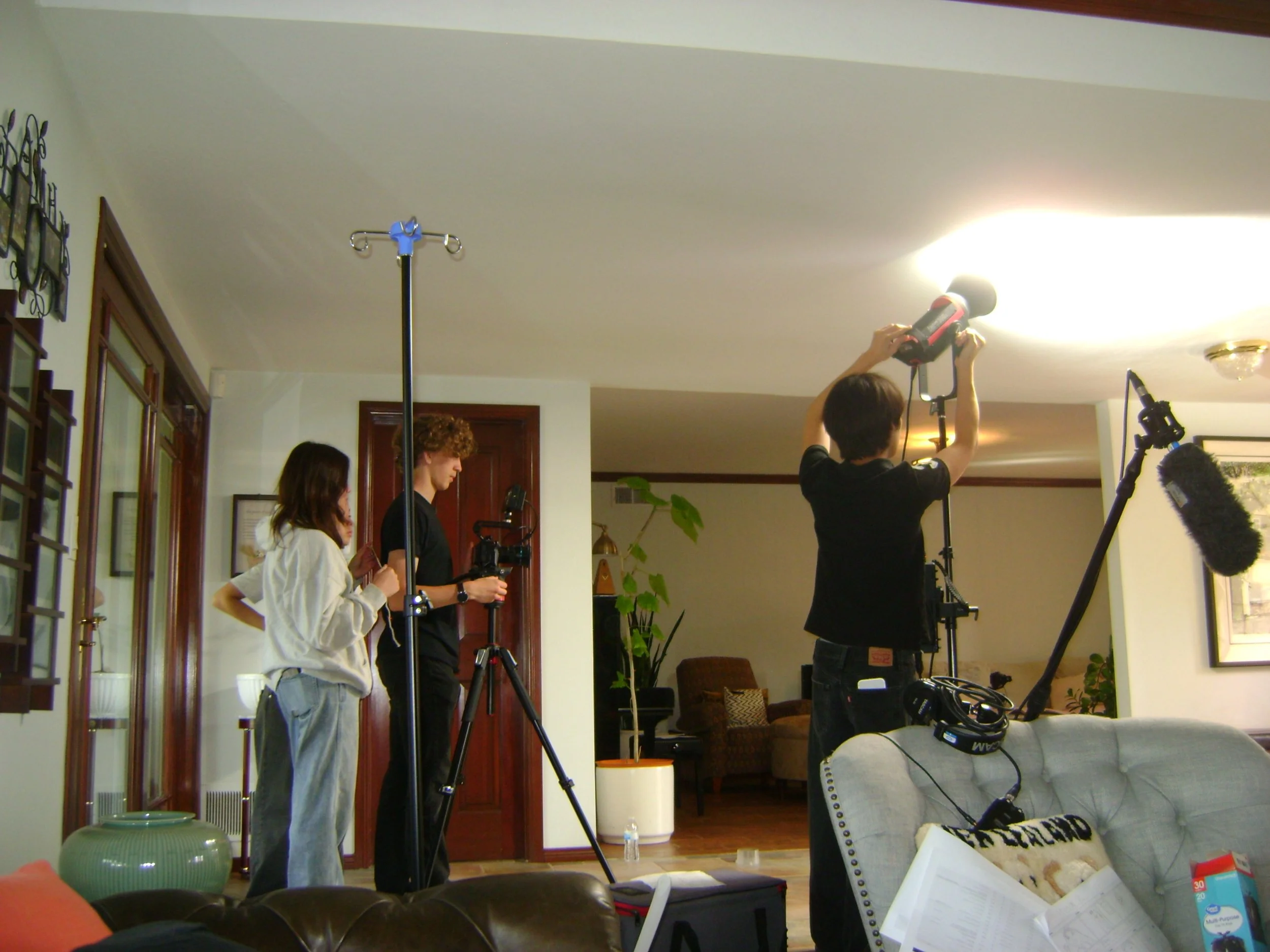 Film crew shooting a scene indoors with cameras, lighting equipment, and a boom microphone in a living room