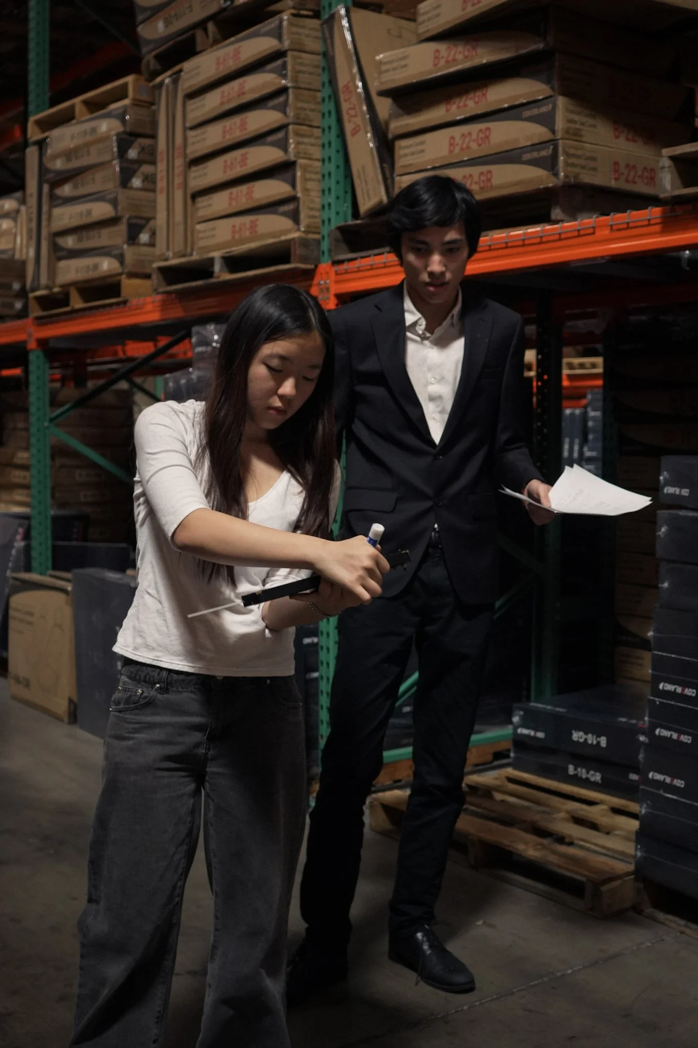 Two people inspecting inventory in a warehouse aisle surrounded by stacked cardboard boxes and shelving.