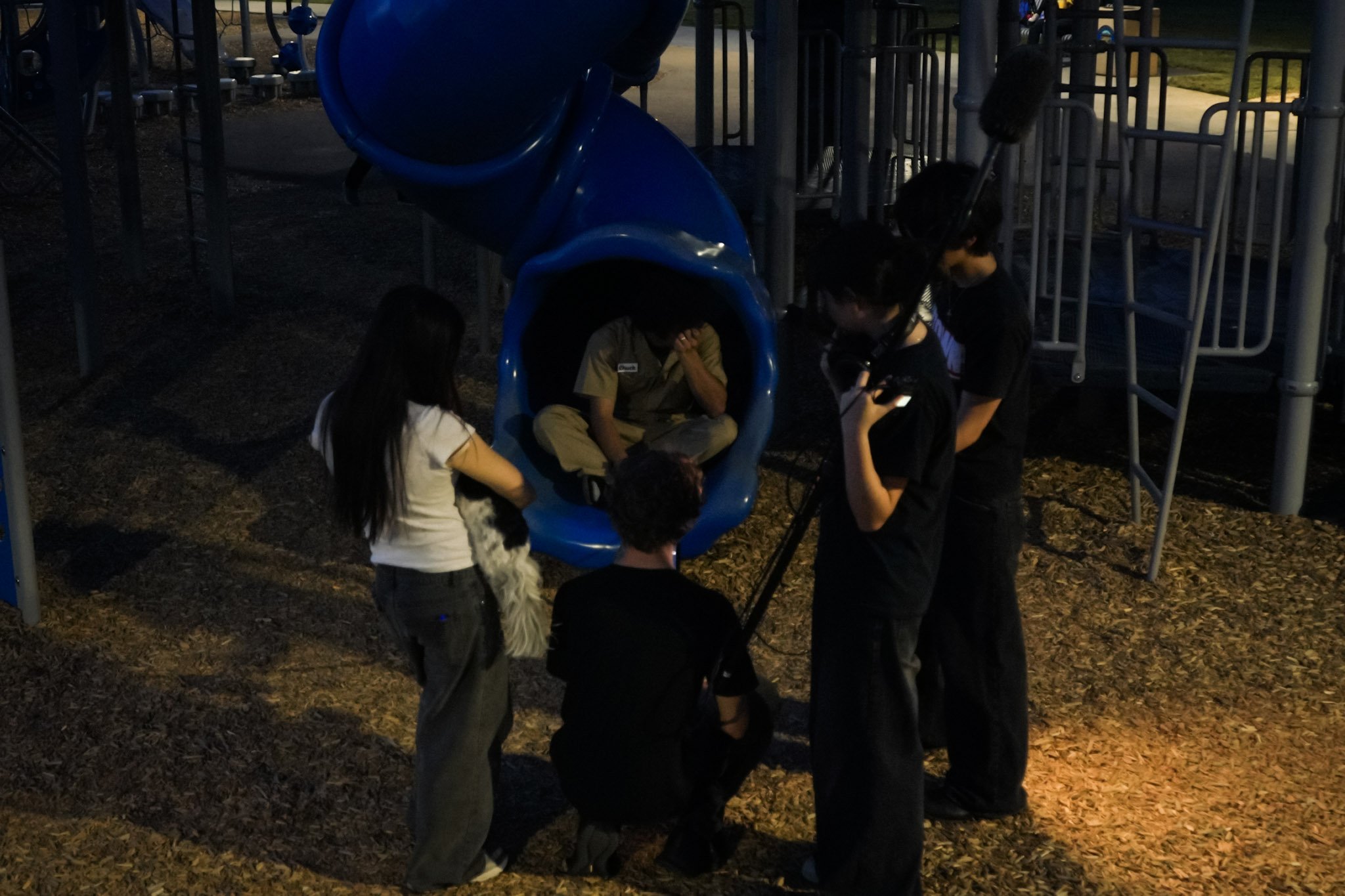 Group of people gathered around a child sitting inside a blue playground tube slide at dusk