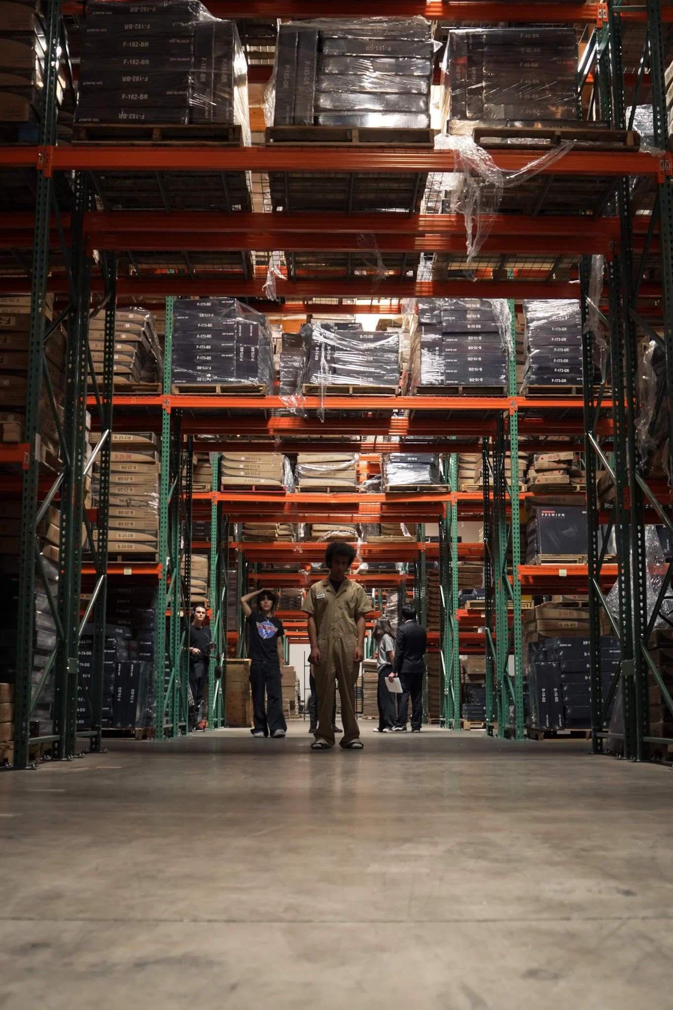 People standing in a tall warehouse aisle surrounded by pallet racks packed with wrapped boxes.