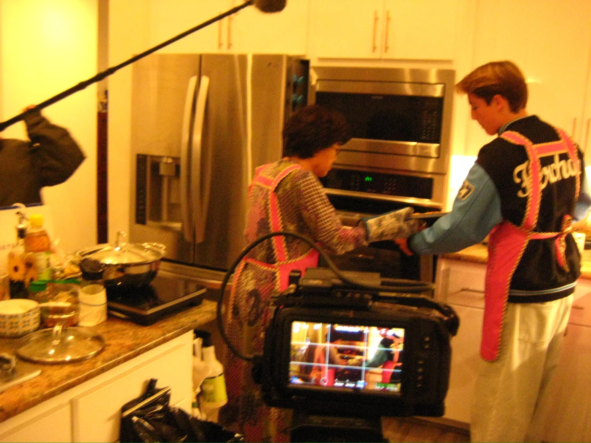 Two people cook in a home kitchen while a camera monitor records the scene.