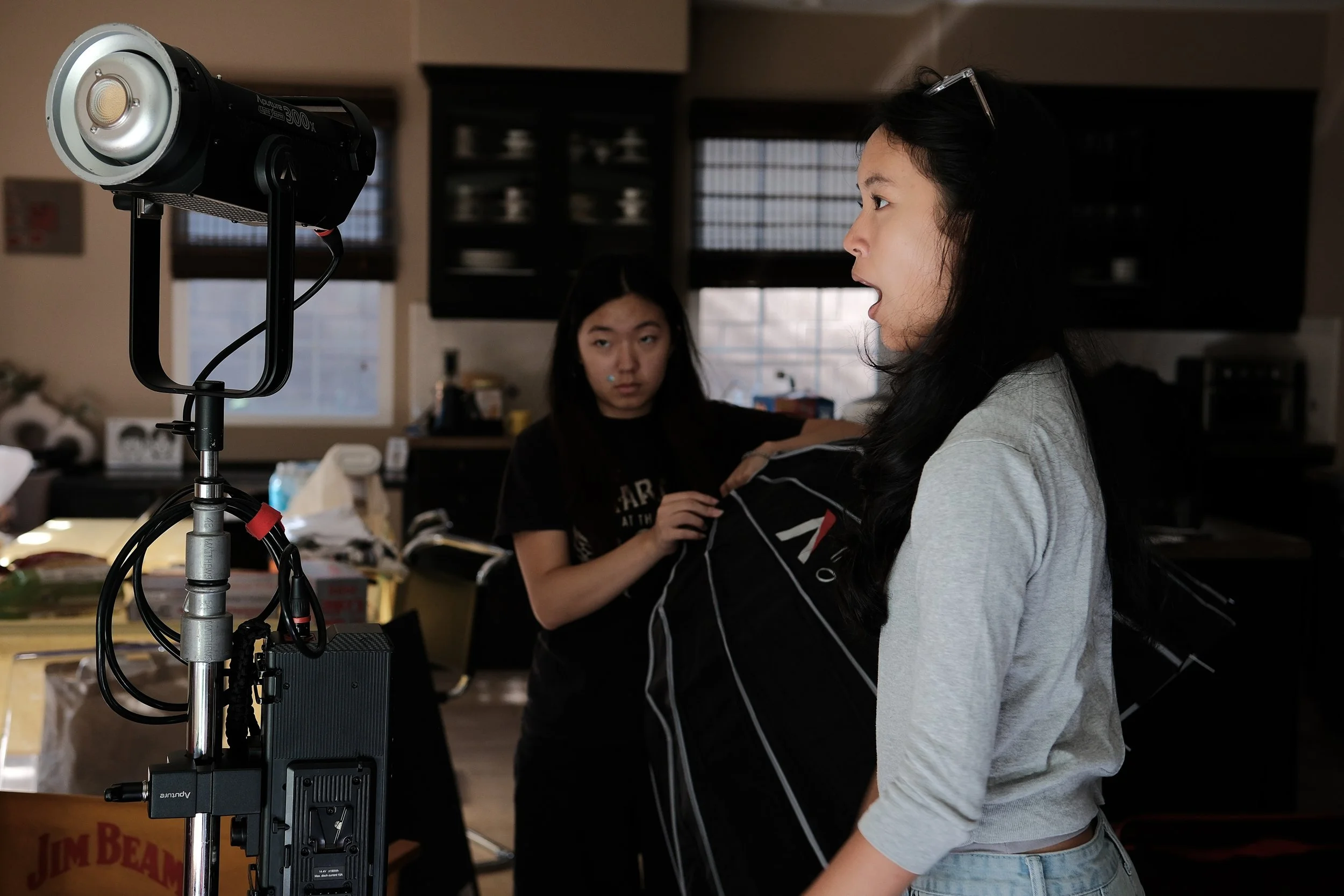 Film crew members adjust lighting equipment while a woman stands in profile on set.
