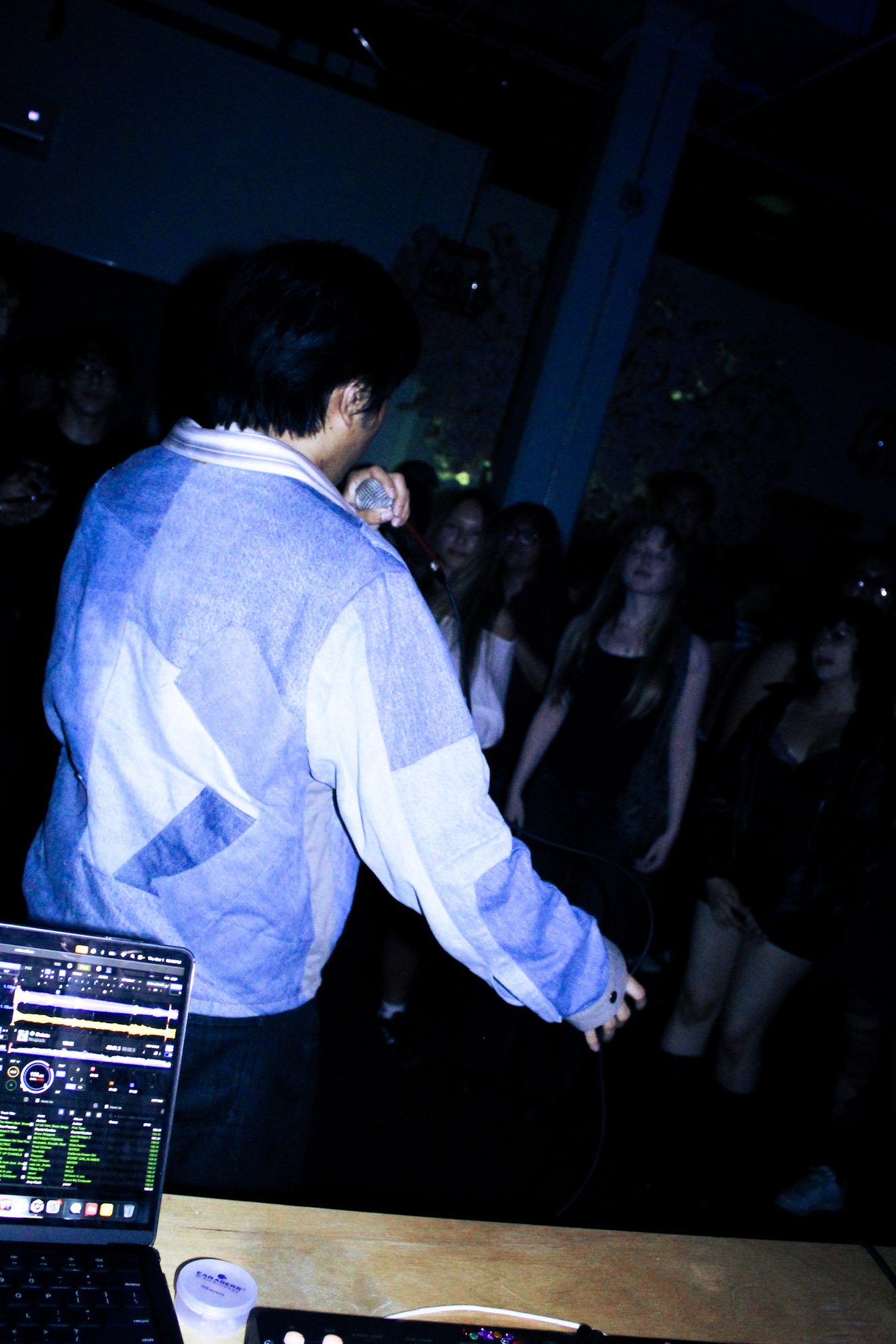 Singer performing into a microphone for a crowd at a dark nightclub, with a laptop DJ setup in the foreground.