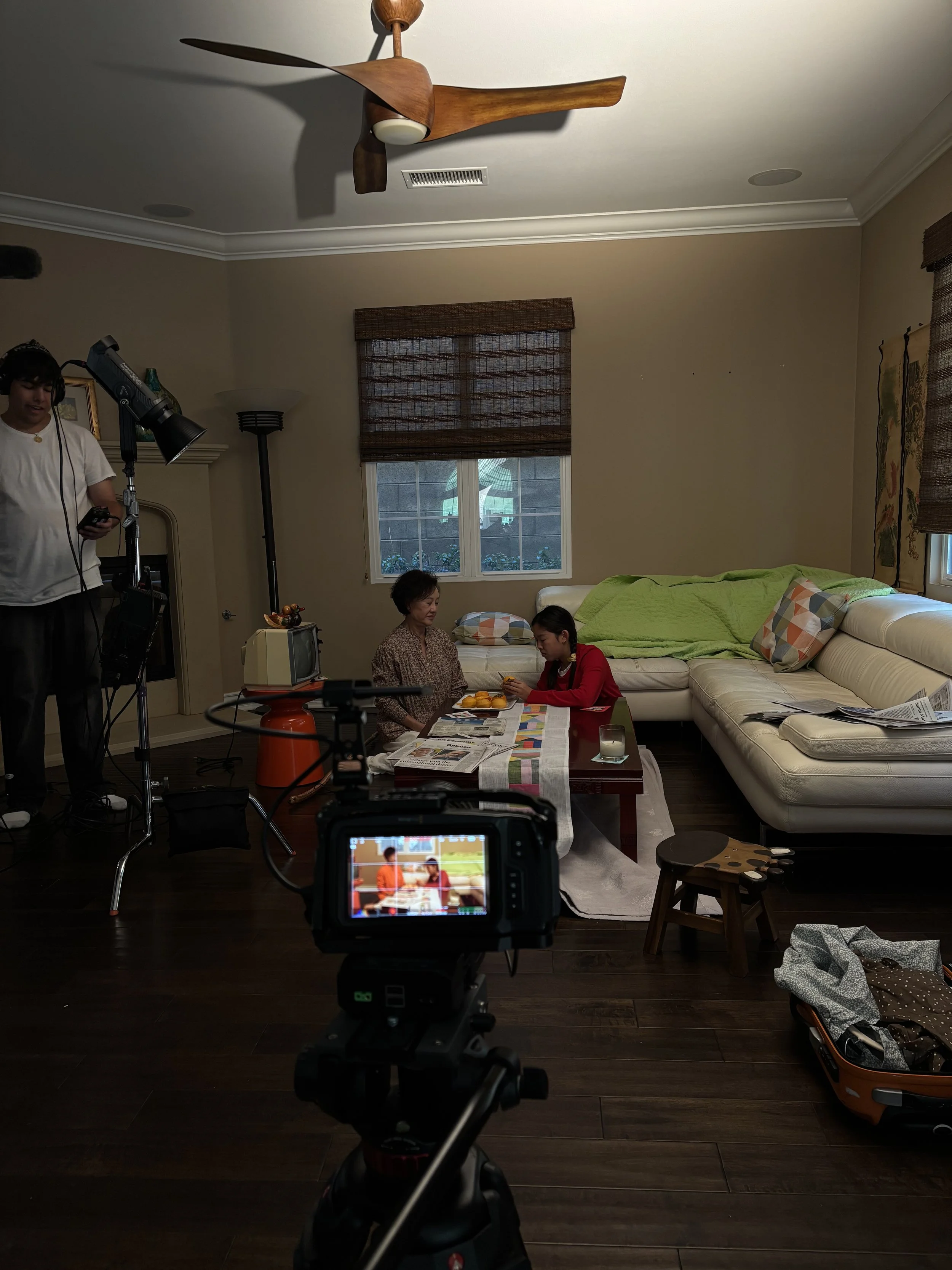 Film crew recording two people eating fruit in a living room with a ceiling fan, camera, lighting equipment, and sofa.