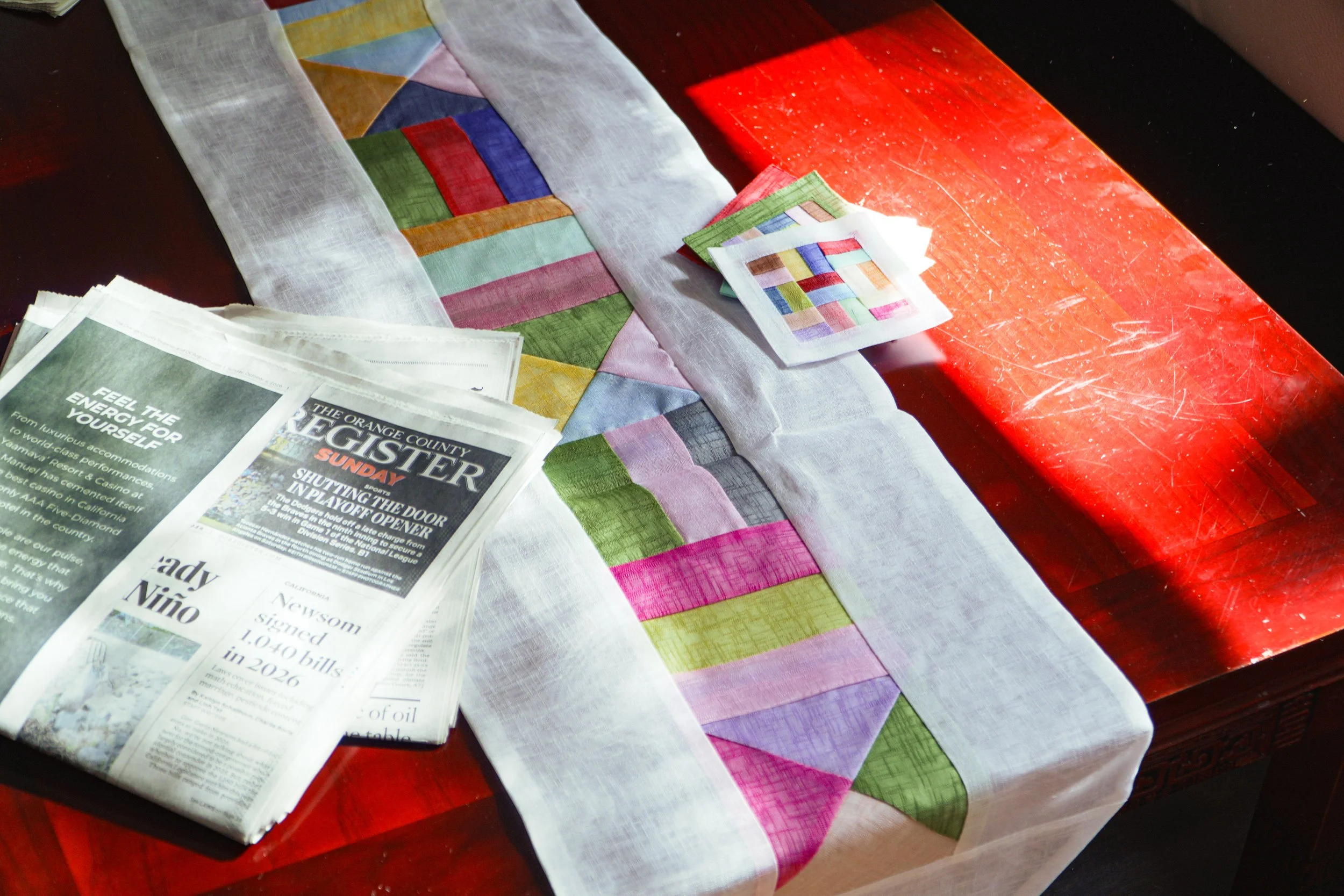 Colorful patchwork quilt and fabric samples spread across a red wooden table beside an open newspaper.
