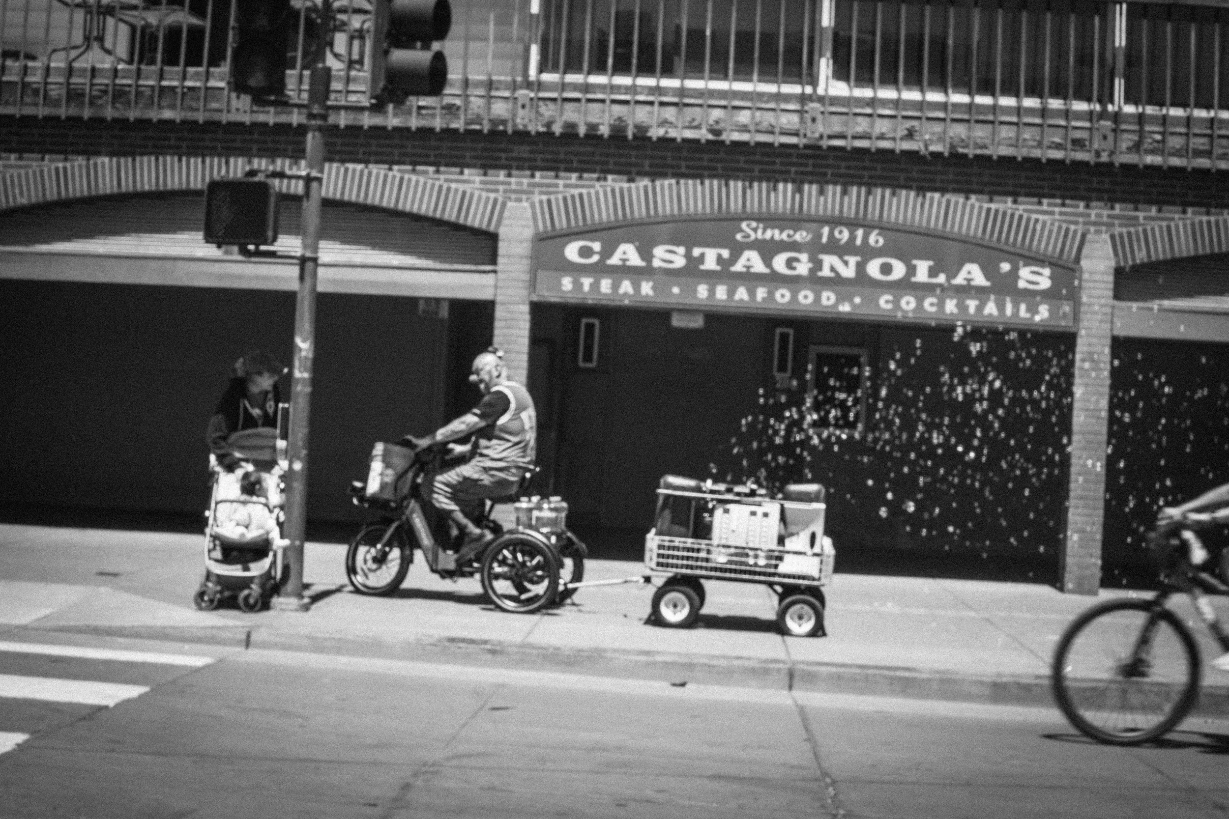 Black-and-white street scene outside Castagnola's steak and seafood restaurant, with a wheelchair user, stroller, cart, and cyclist.