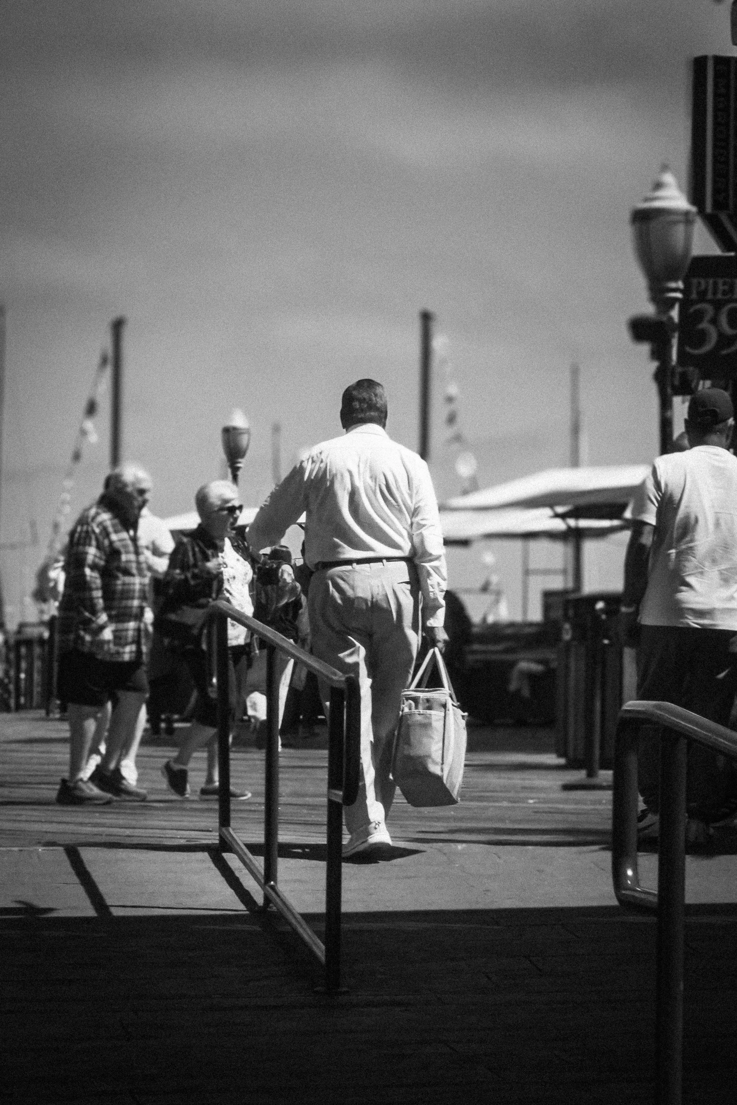 Black-and-white street scene with pedestrians walking past outdoor café seating and a man in a white shirt carrying a shopping bag.