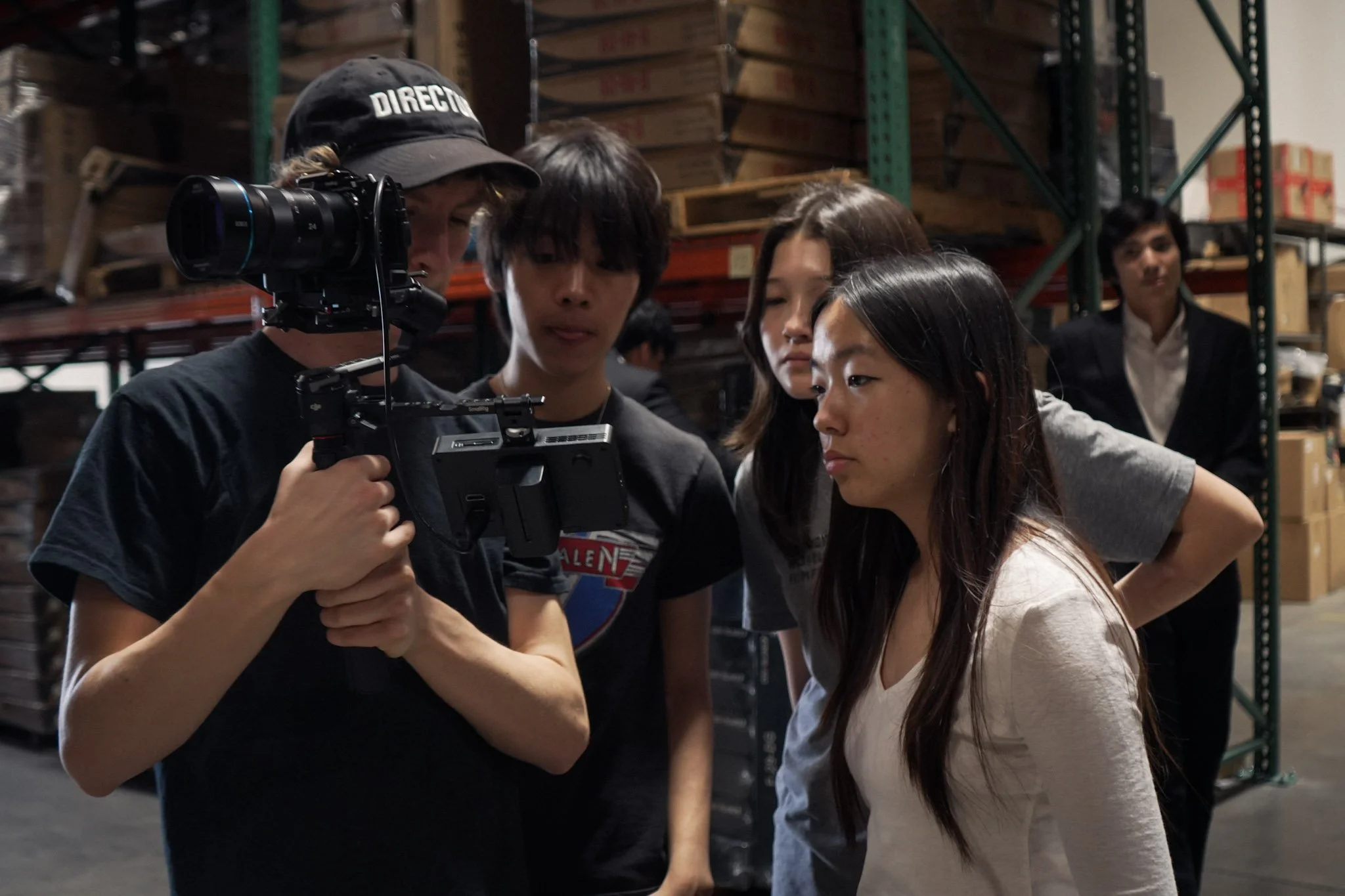 Film crew reviewing camera footage inside a warehouse surrounded by stacked crates