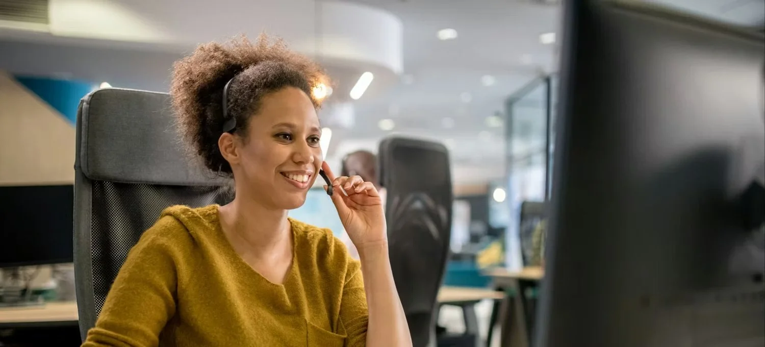 A woman wearing a mustard yellow sweater and a headset is smiling while working at a computer in an office setting.